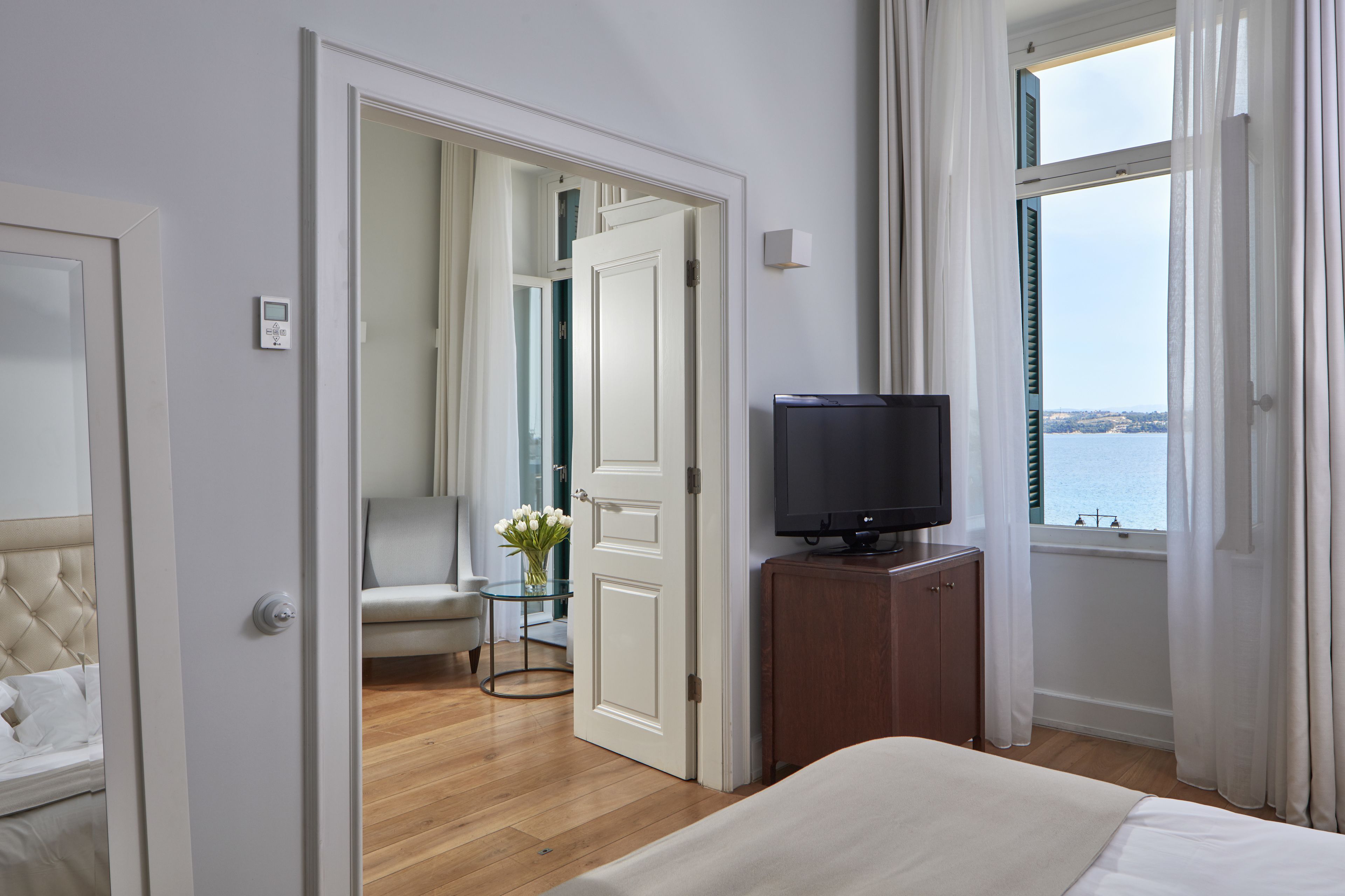 A hotel room with a bed, a television on a wooden stand, a small table with a vase of flowers, and a view of the sea through a window.