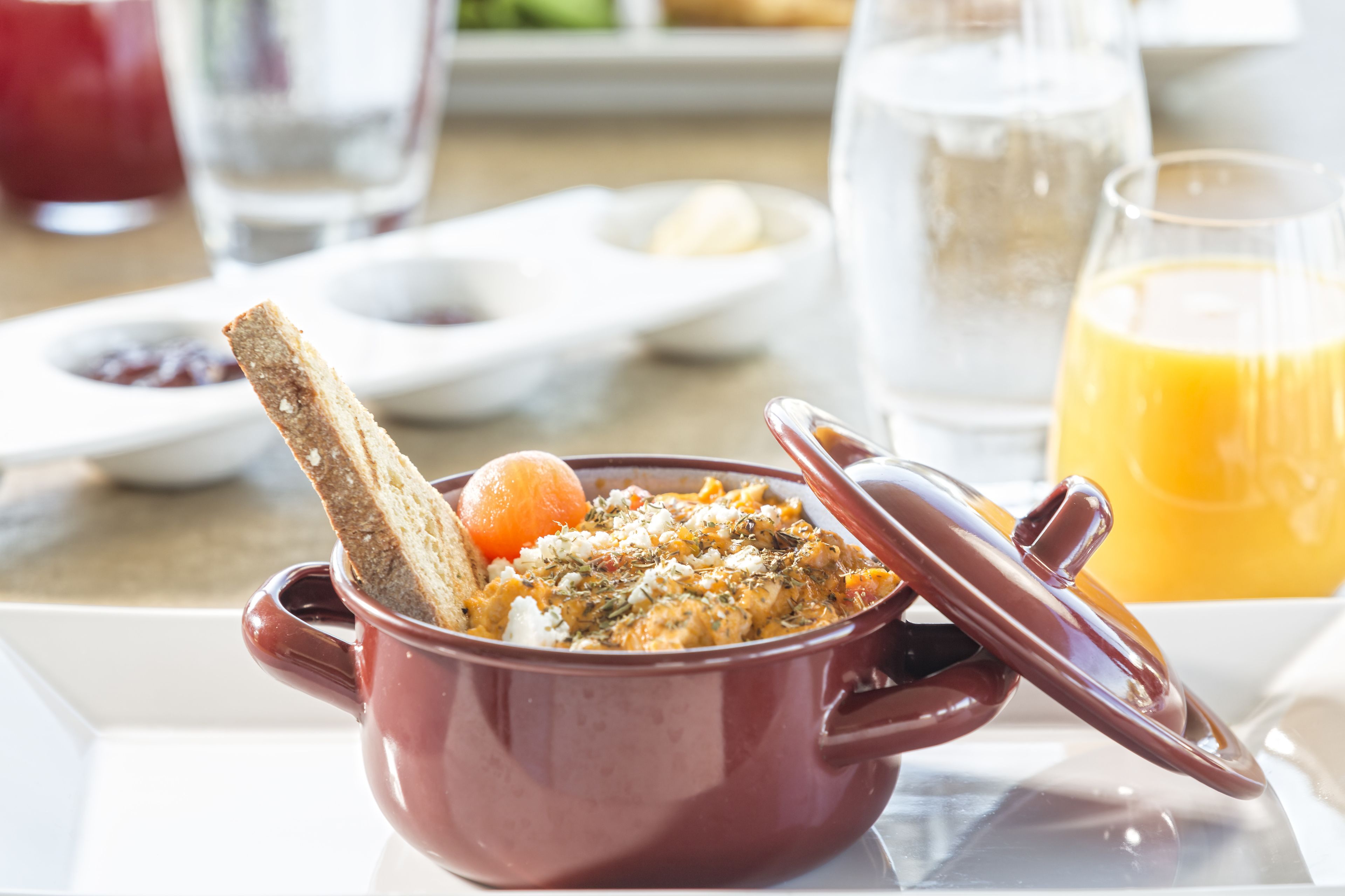 A breakfast setting with a red pot containing a dish, a glass of orange juice, and a plate with toast and a boiled egg.