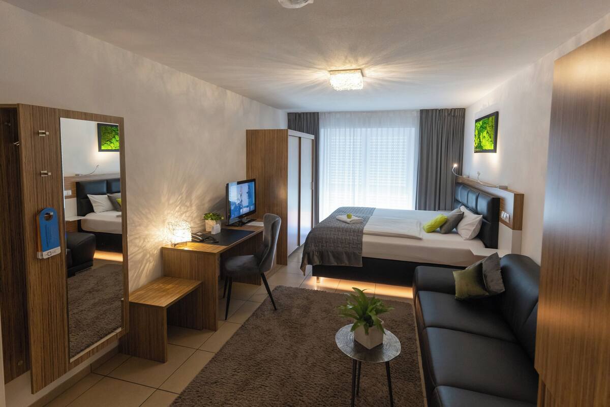 Ein modernes Hotelzimmer mit Bett, Schreibtisch, Stuhl, Sofa, Fernseher und einem kleinen Tisch mit Pflanze.