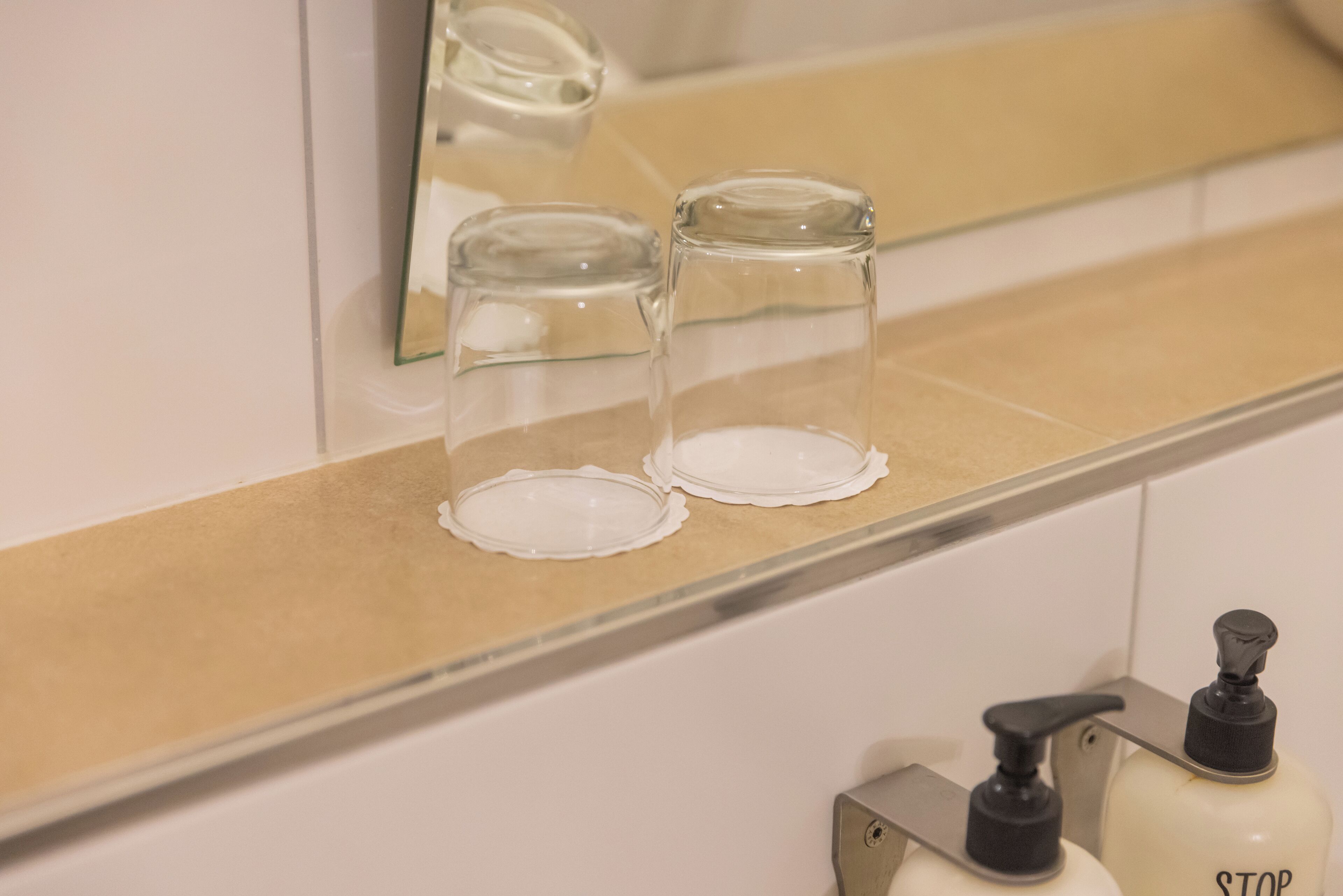 Un baño con un espejo, dos recipientes de vidrio transparente sobre una encimera color beige y un dispensador de jabón con la etiqueta 'STOP' en un soporte metálico.