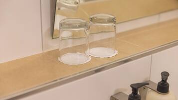Un baño con un espejo, dos recipientes de vidrio transparente sobre una encimera color beige y un dispensador de jabón con la etiqueta 'STOP' en un soporte metálico.