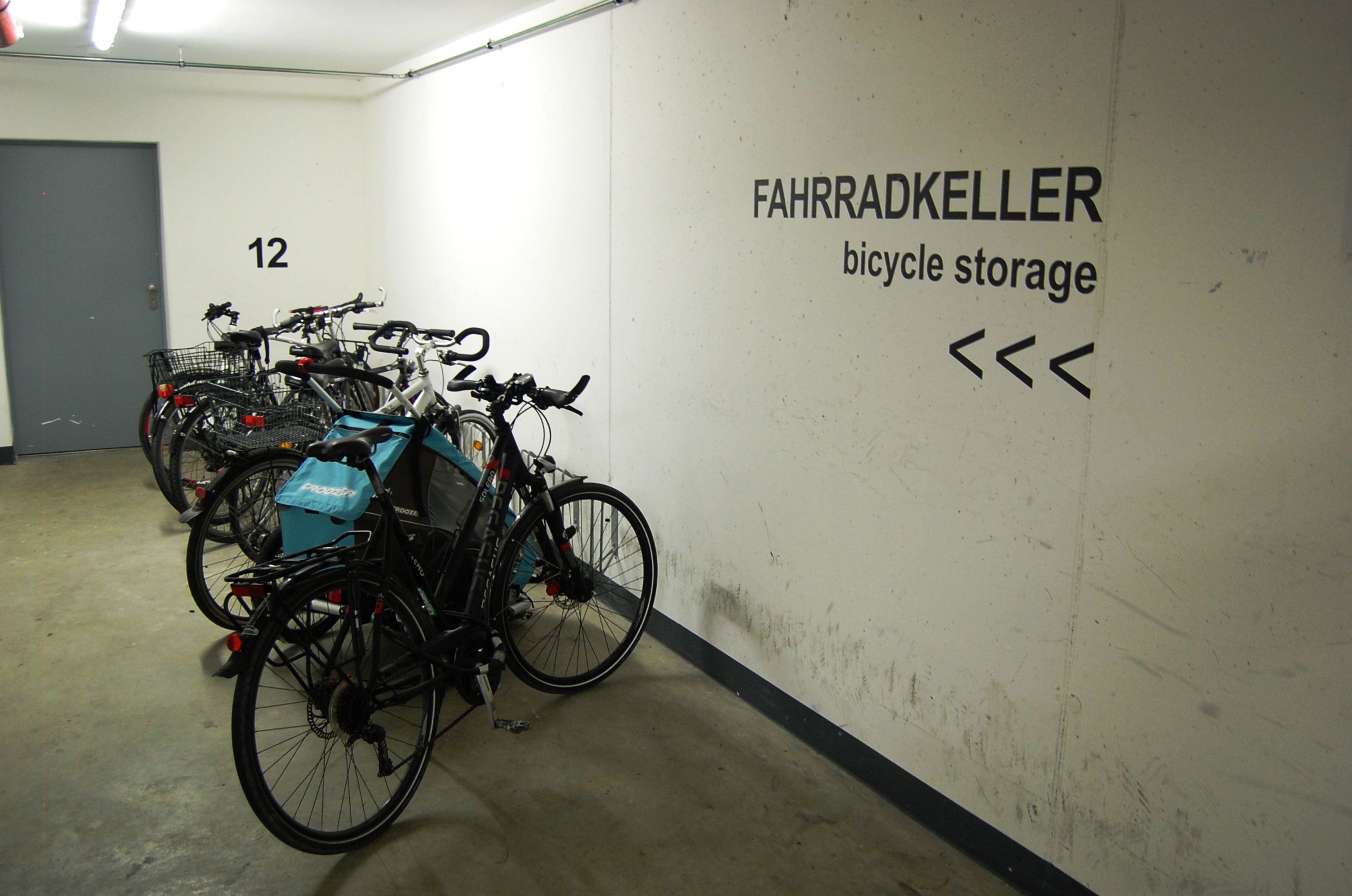 Ein Fahrradabstellraum mit mehreren Fahrrädern, die an der Wand lehnen. Beschriftung: FAHRRADKELLER Fahrradabstellraum.