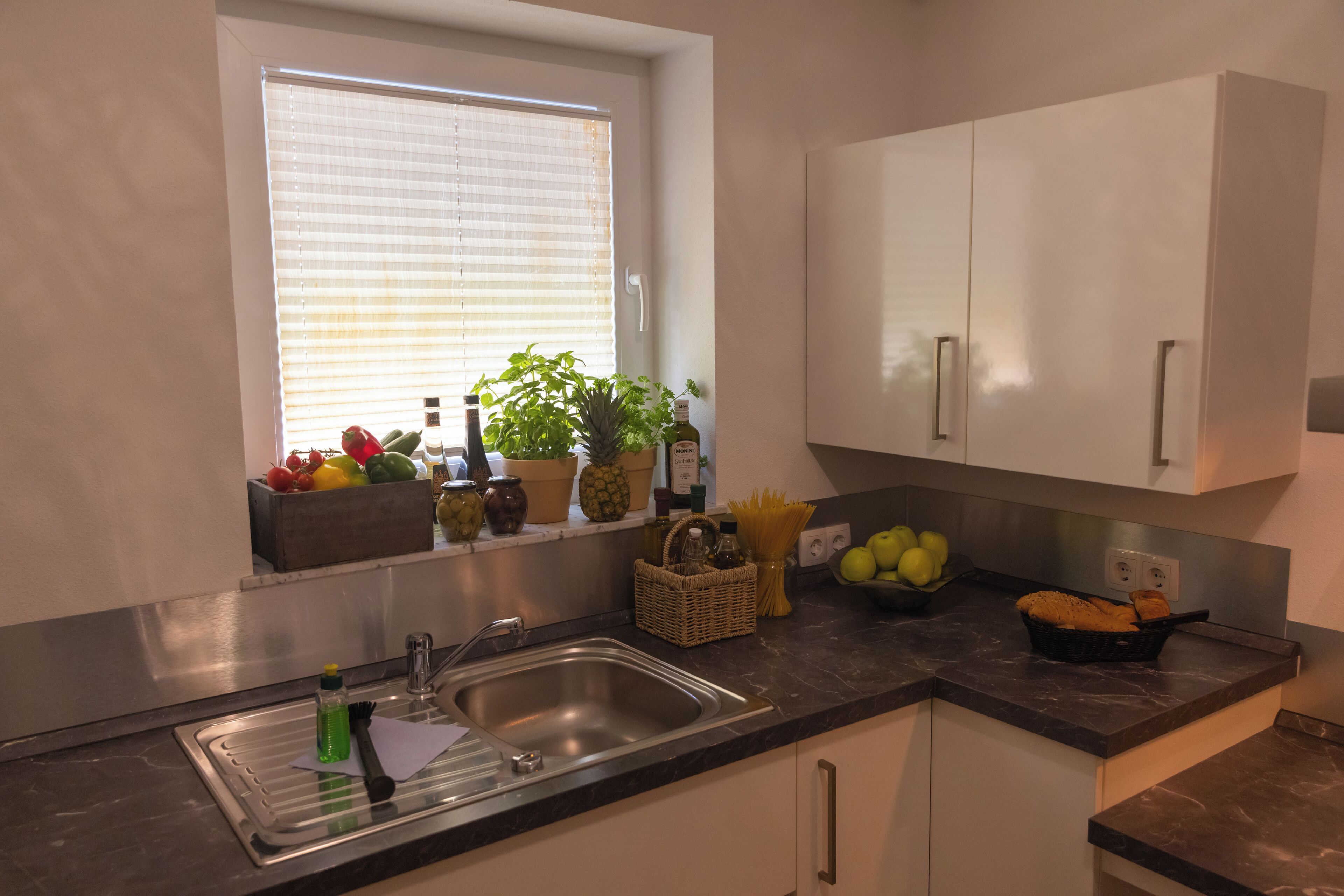 Una cocina con ventana, pileta de acero inoxidable, muebles blancos y varios utensilios de cocina sobre la encimera.