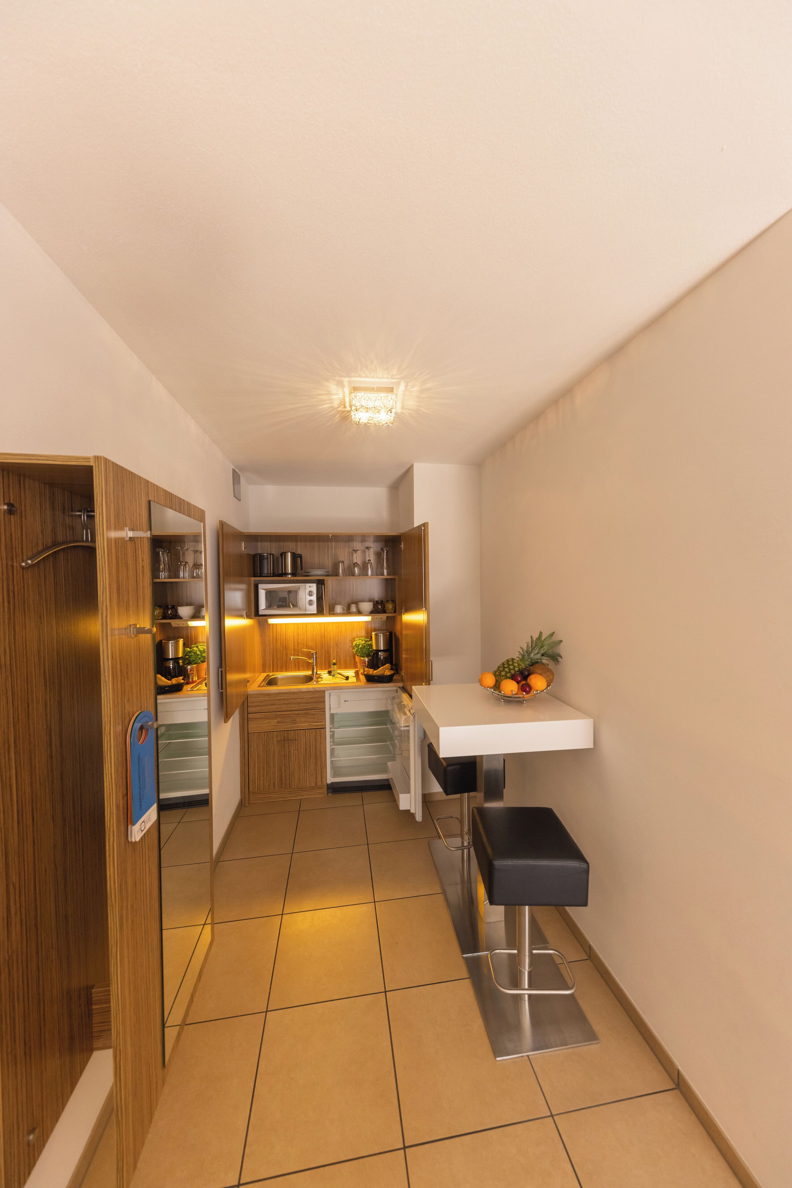 A narrow, well-lit hallway leading to a kitchen with wooden cabinets, a microwave, and a fruit bowl on a white counter.