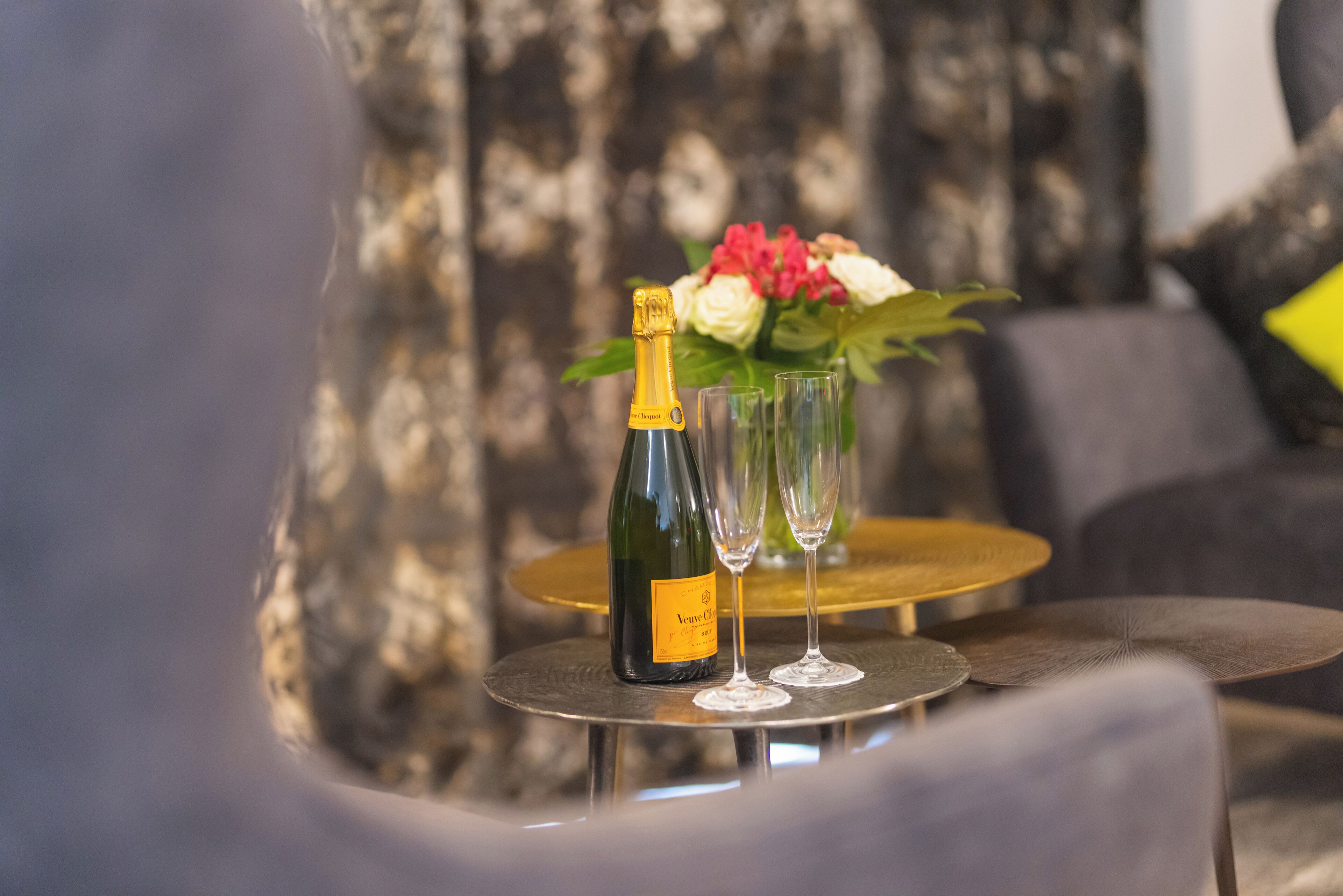 A bottle of champagne, two glasses, and a vase with flowers on a round table.