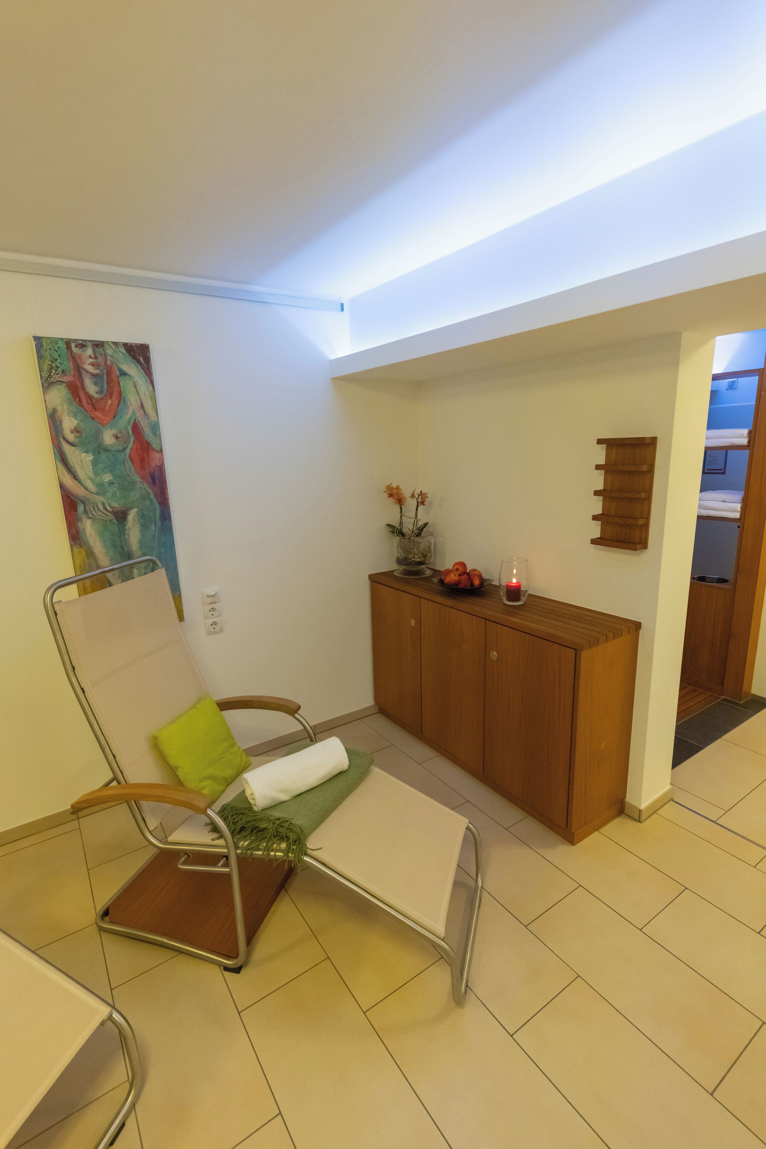 Una habitación con un aparador de madera, un cojín verde en una silla metálica, un florero con flores y una vela sobre el aparador.