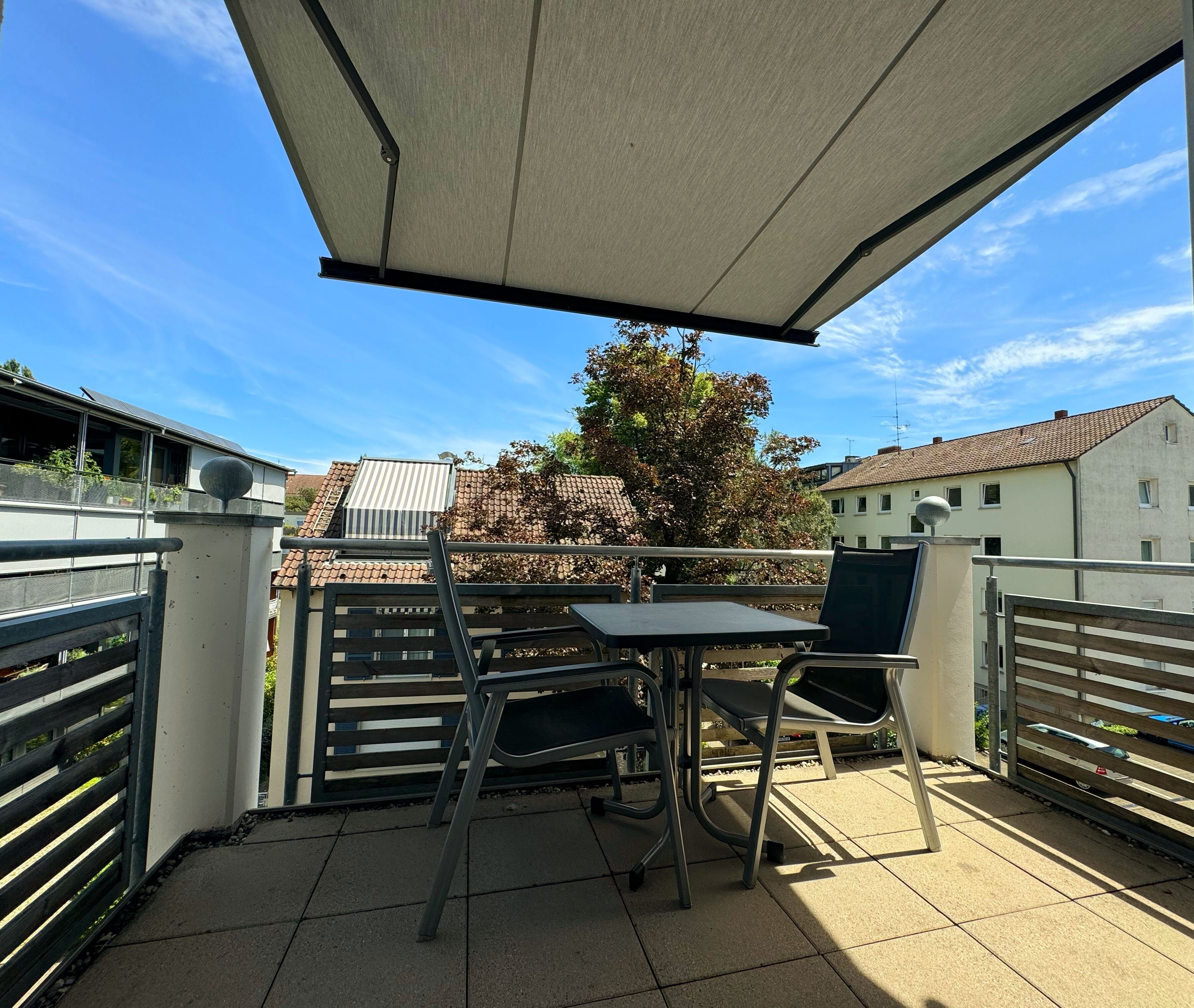Ein Balkon mit Tisch und Stühlen, einem Metallgeländer und Blick auf Gebäude und Bäume.