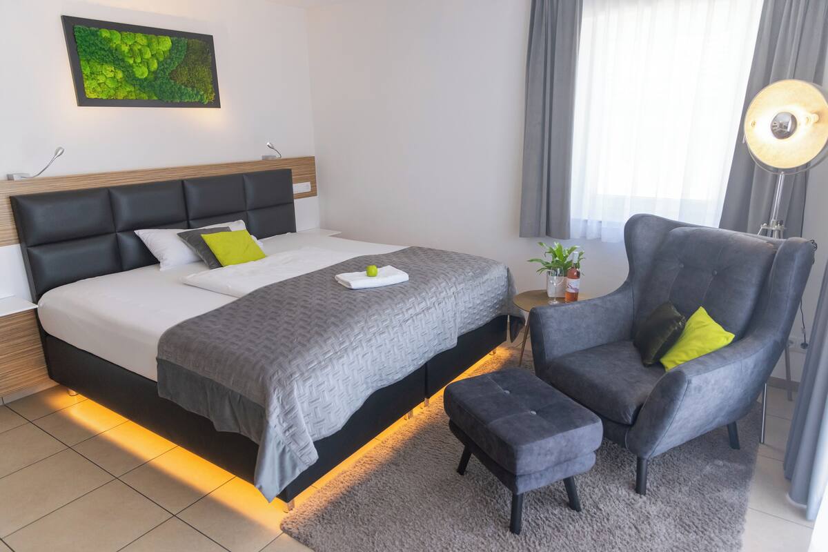 Ein modernes Hotelzimmer mit einem großen Bett, einem grauen Sessel, einem kleinen Tisch mit Pflanze und einer Nachttischlampe.
