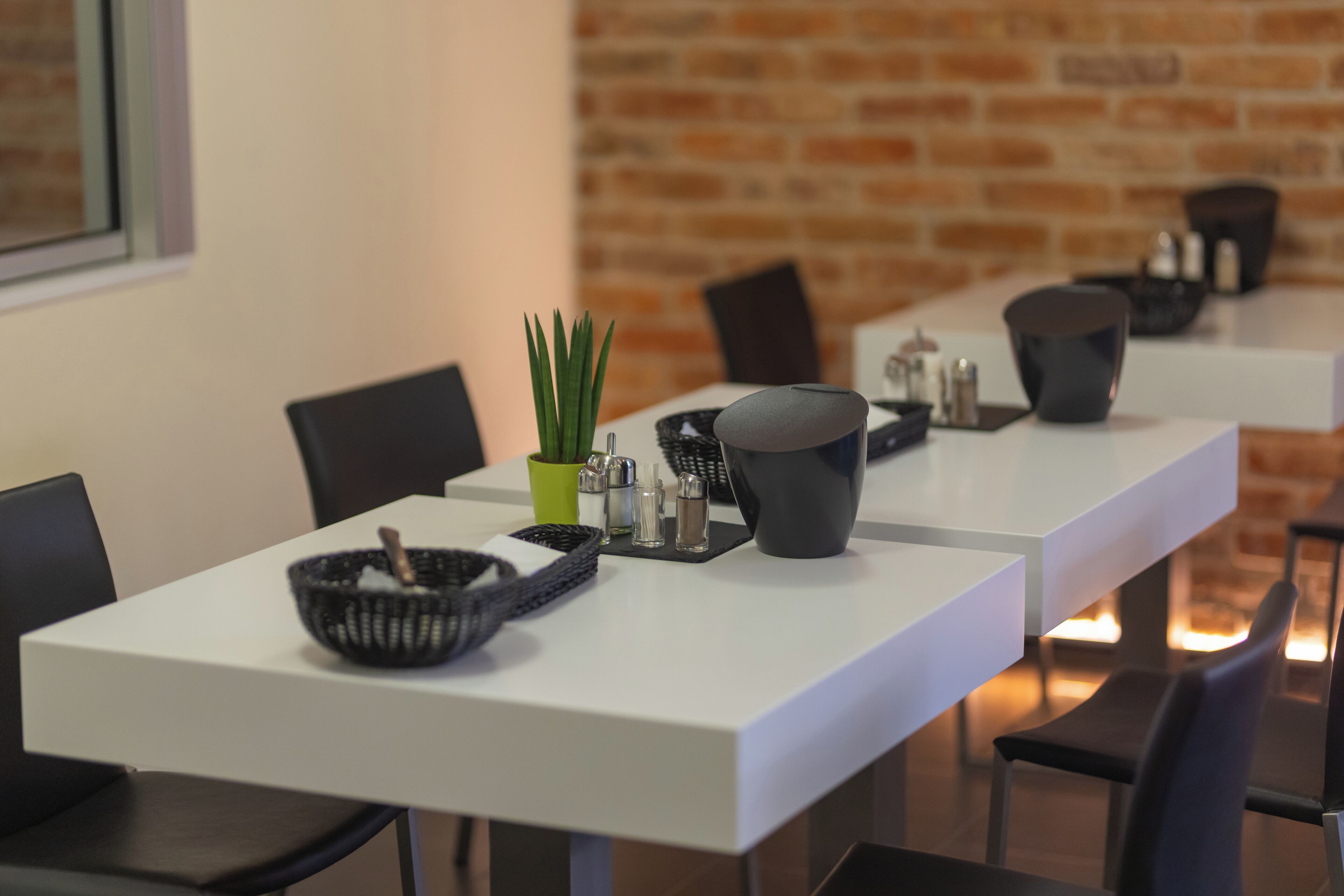 Un comedor moderno con mesas blancas, sillas negras y una pared de ladrillo al fondo.
