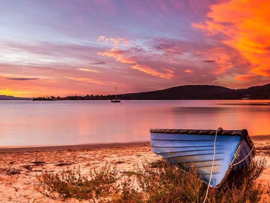 Un bote en una playa arenosa con una masa de agua tranquila y una puesta de sol al fondo.