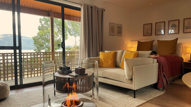 A modern living room with a fireplace, a sofa with yellow cushions, and a view of the outdoors. - Driftwood Cottages (Dover)