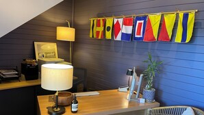 A hotel room with a wooden desk, a lamp, a laptop, a bottle of wine, a potted plant, and a chair with a cushion. Flags are hanging on the wall. - Driftwood Cottages (Dover)