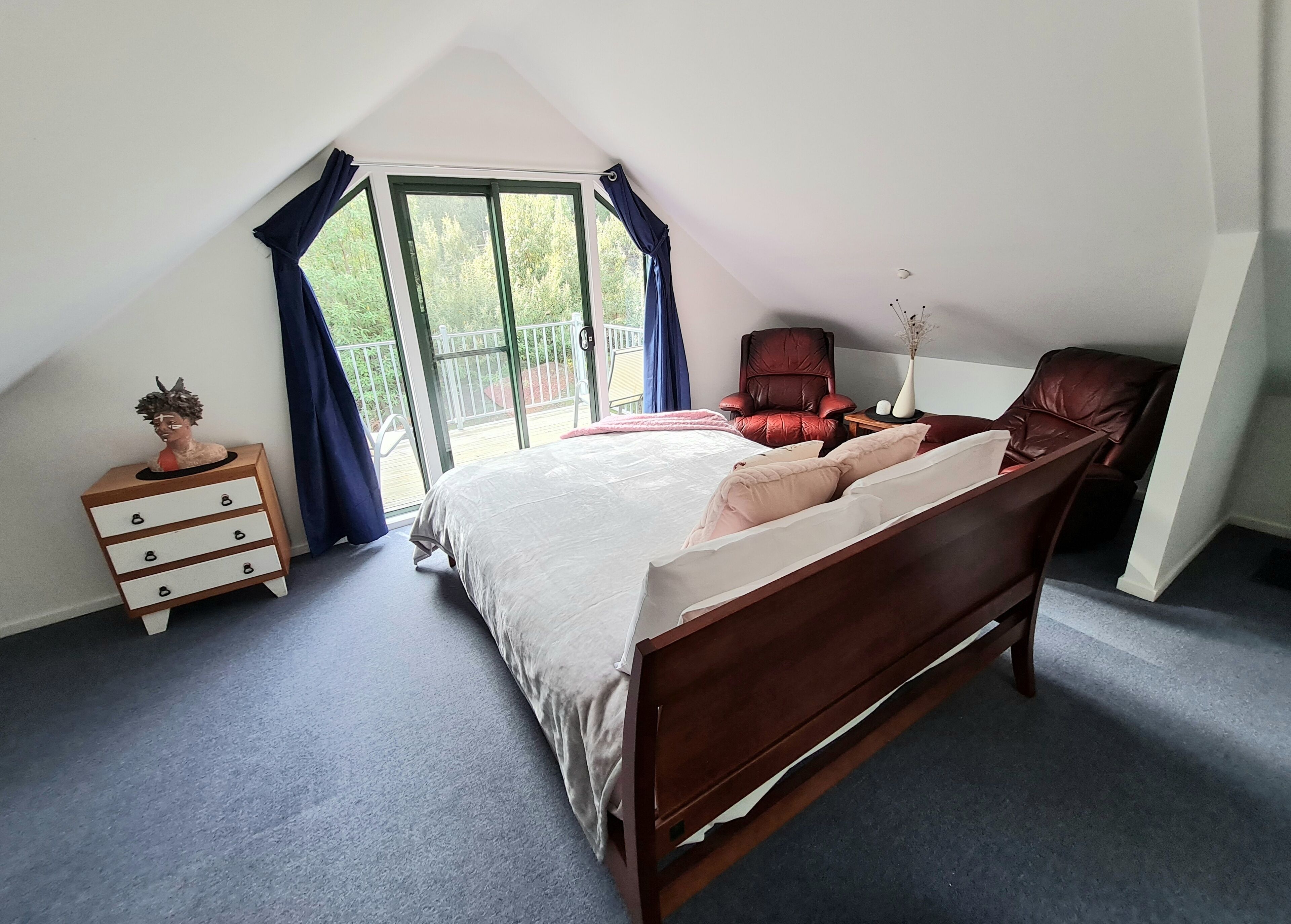 Een slaapkamer met een schuine kap, een groot bed met houten frame, een nachtkastje met lades, een rode stoel en uitzicht door een glazen deur.