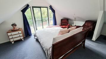 Een slaapkamer met een schuine kap, een groot bed met houten frame, een nachtkastje met lades, een rode stoel en uitzicht door een glazen deur.