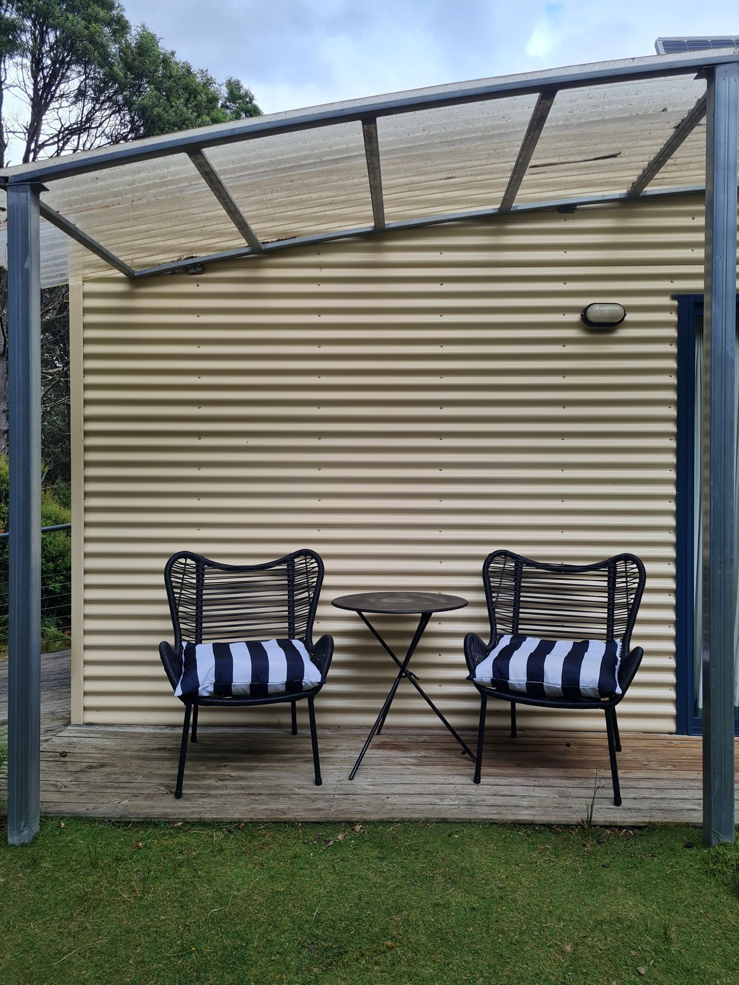 Eine überdachte Terrasse mit zwei schwarzen Korbstühlen und einem kleinen runden Tisch, ausgestattet mit gestreiften Kissen.