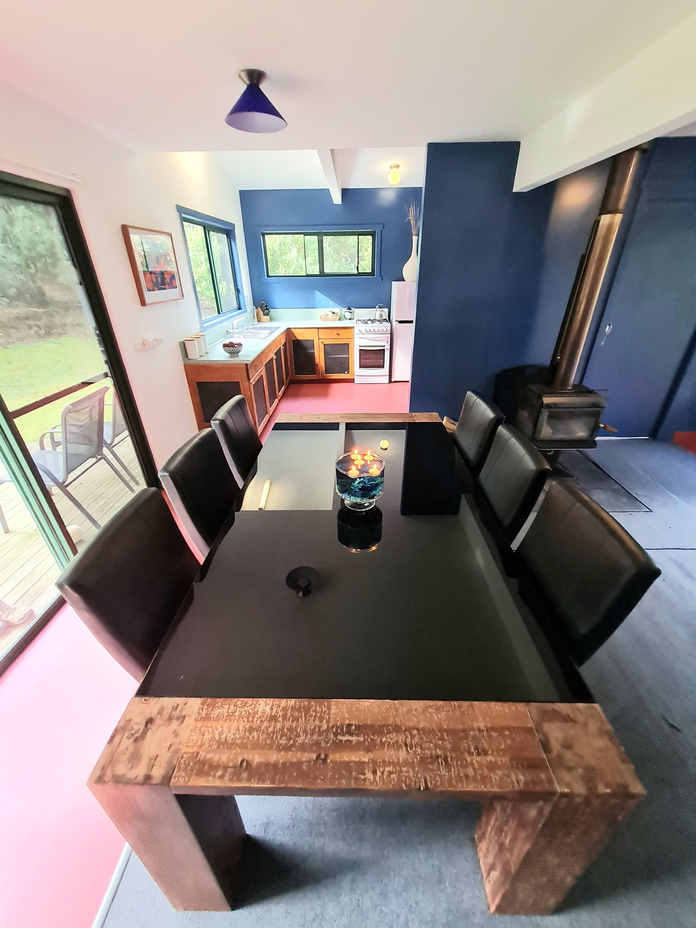 Zona pranzo con tavolo in legno e sedie nere, cucina sullo sfondo e vista sull'esterno.