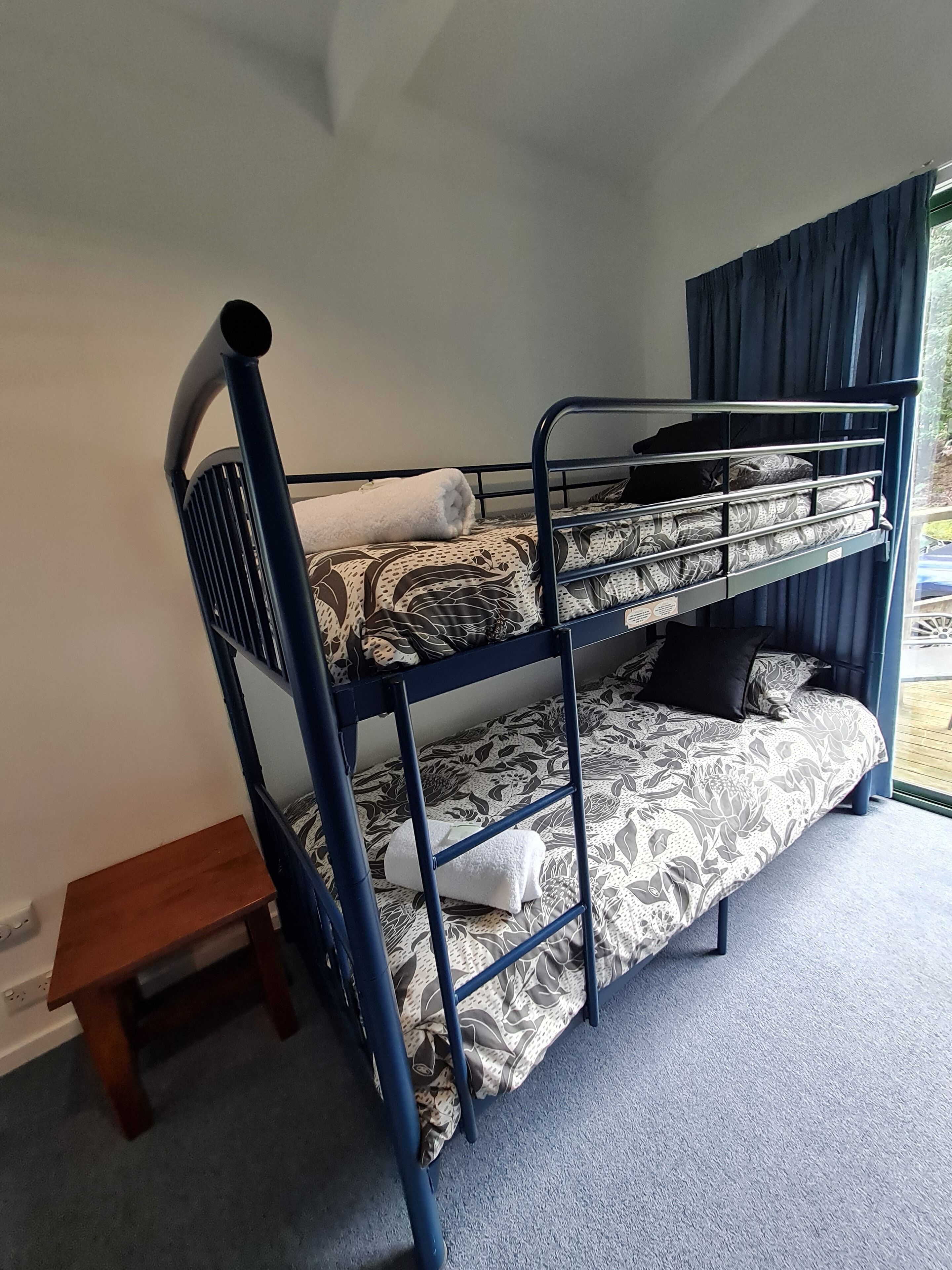 A bunk bed with a patterned bedspread, a small wooden stool, and a window with curtains.