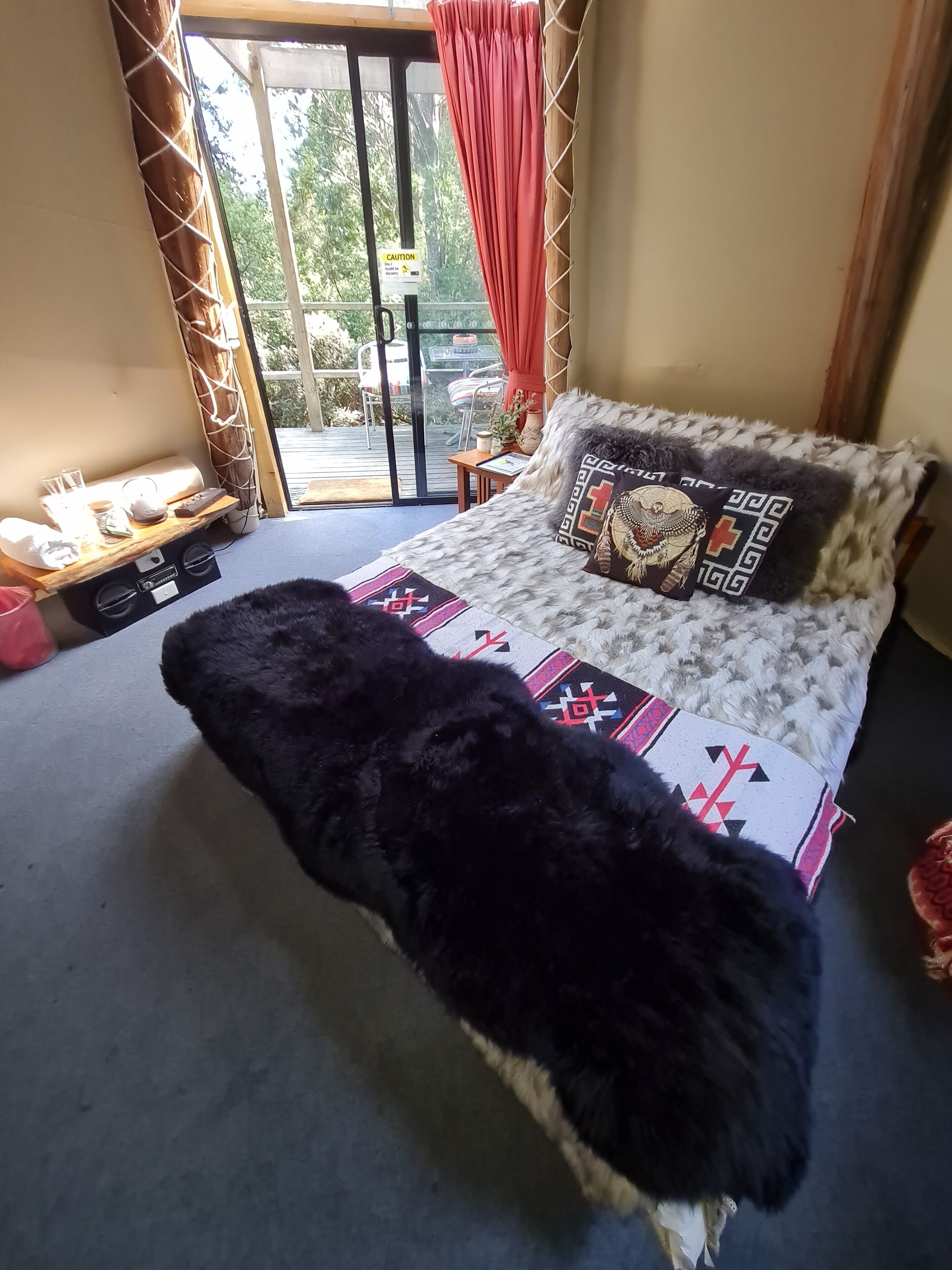 A bedroom with a bed, a window with curtains, a small table with a lamp, and a view of the outdoors.