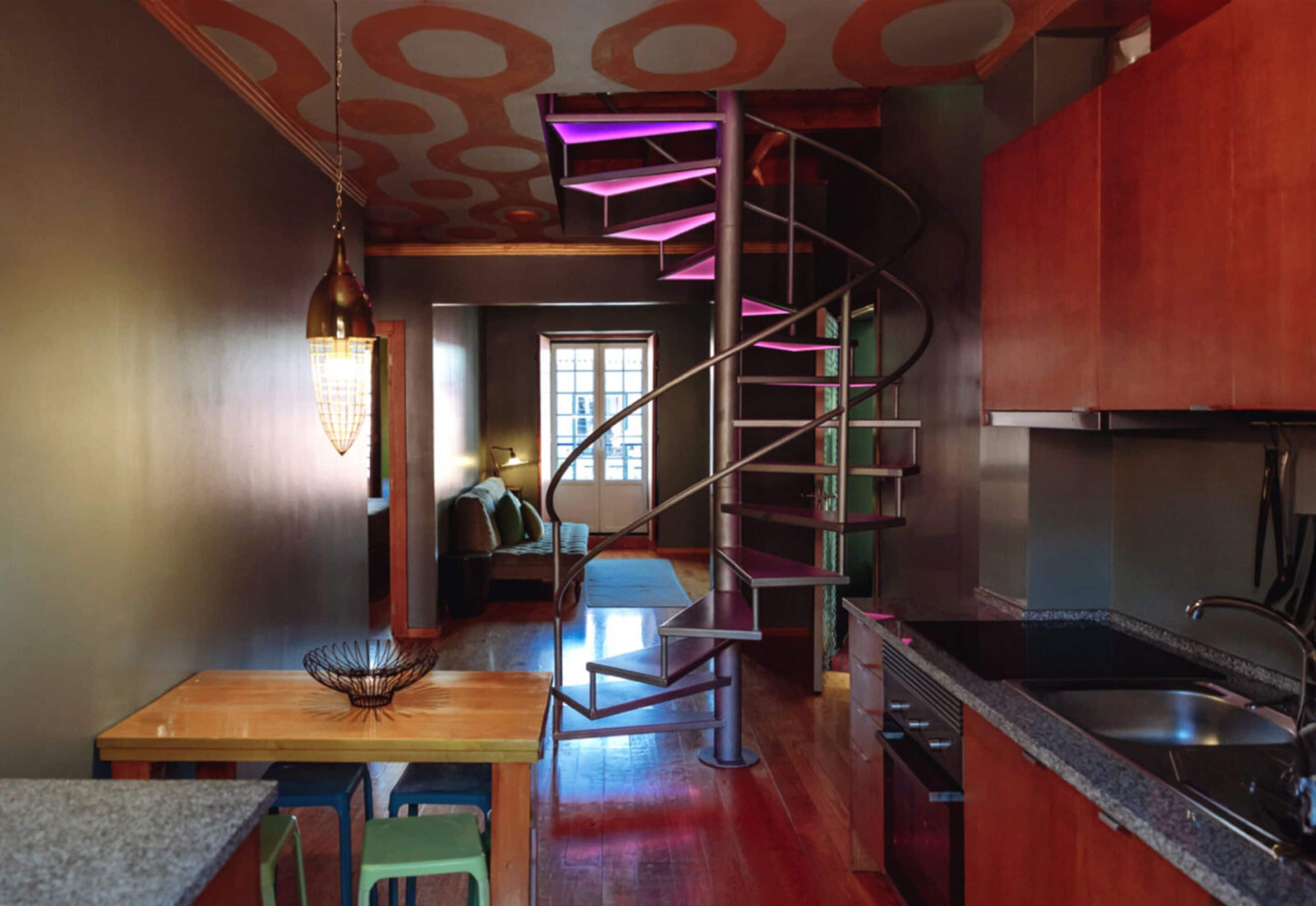A modern kitchen with a spiral staircase, wooden dining table, and a view to the living area.