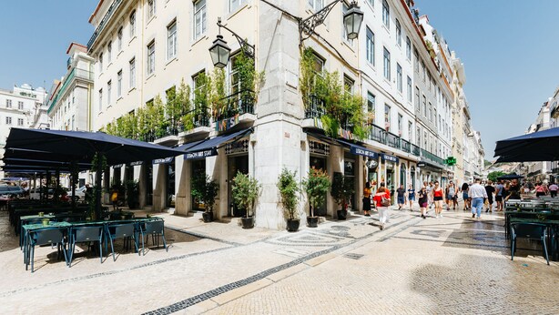 A street with outdoor seating, potted plants, and a building with multiple shops.