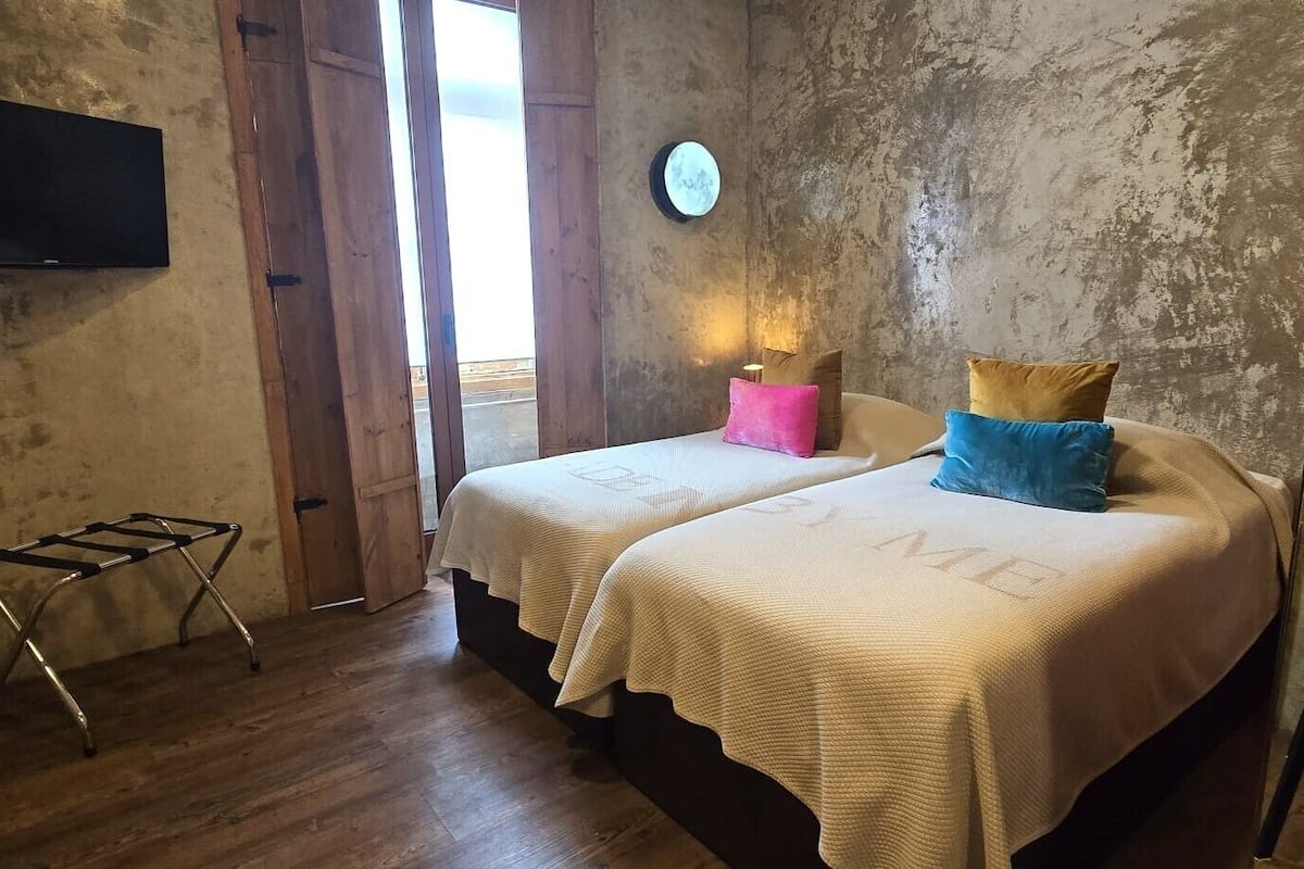Un dormitorio con dos camas, piso de madera, televisor montado en la pared, una mesita y una silla.