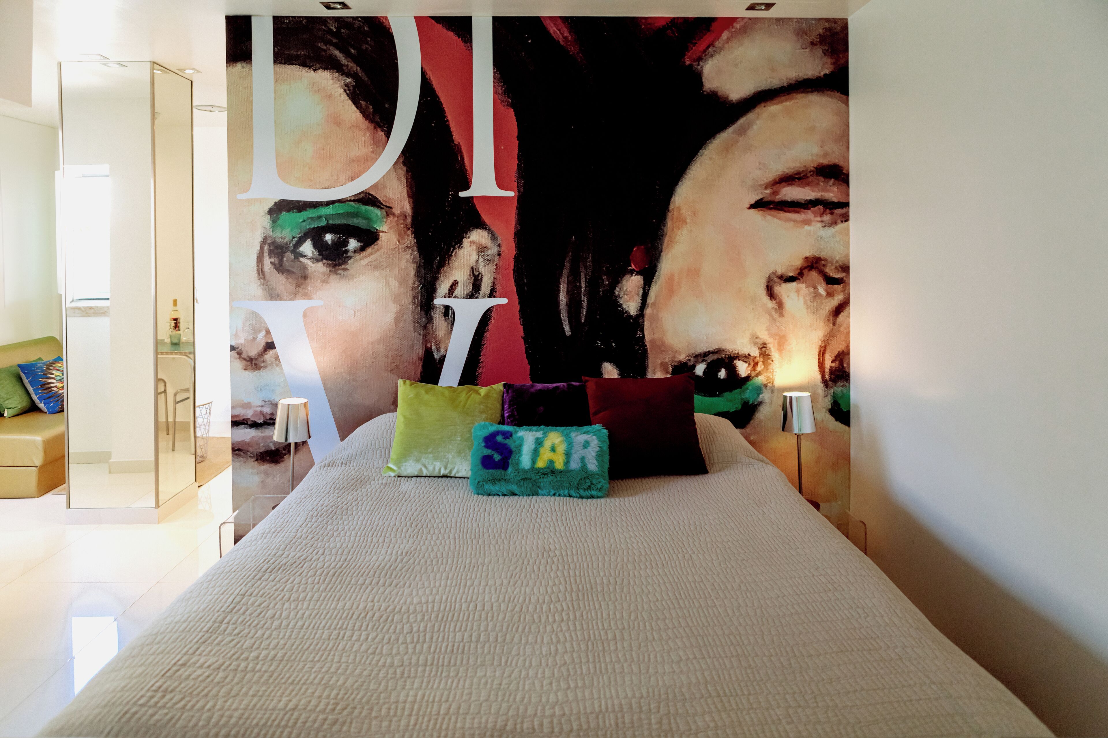 A modern hotel room with a large wall mural, a bed with white bedding, and colorful pillows.