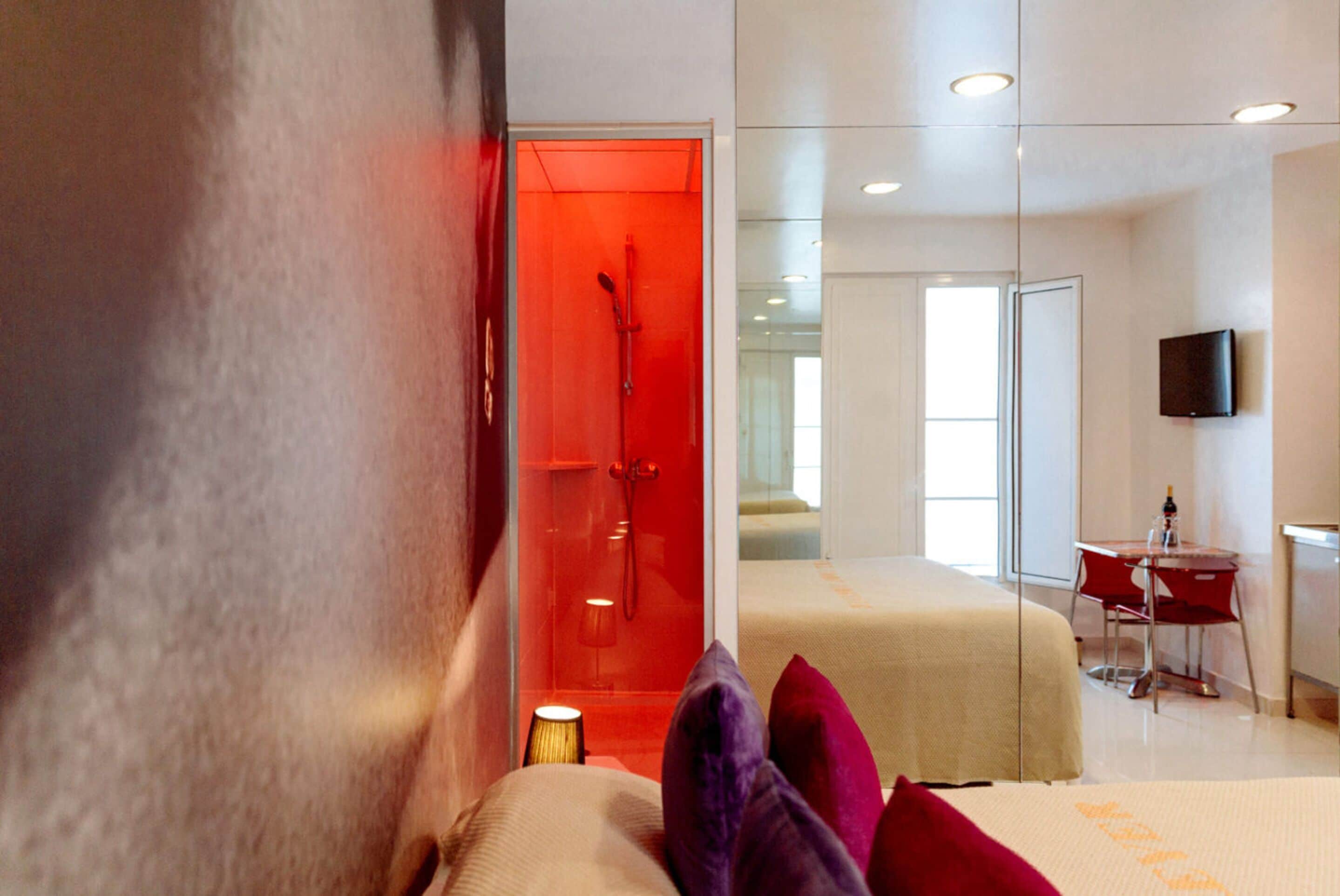 A modern hotel room with a glass shower door, a bed with colorful pillows, a bedside table, and a flat-screen TV mounted on the wall.