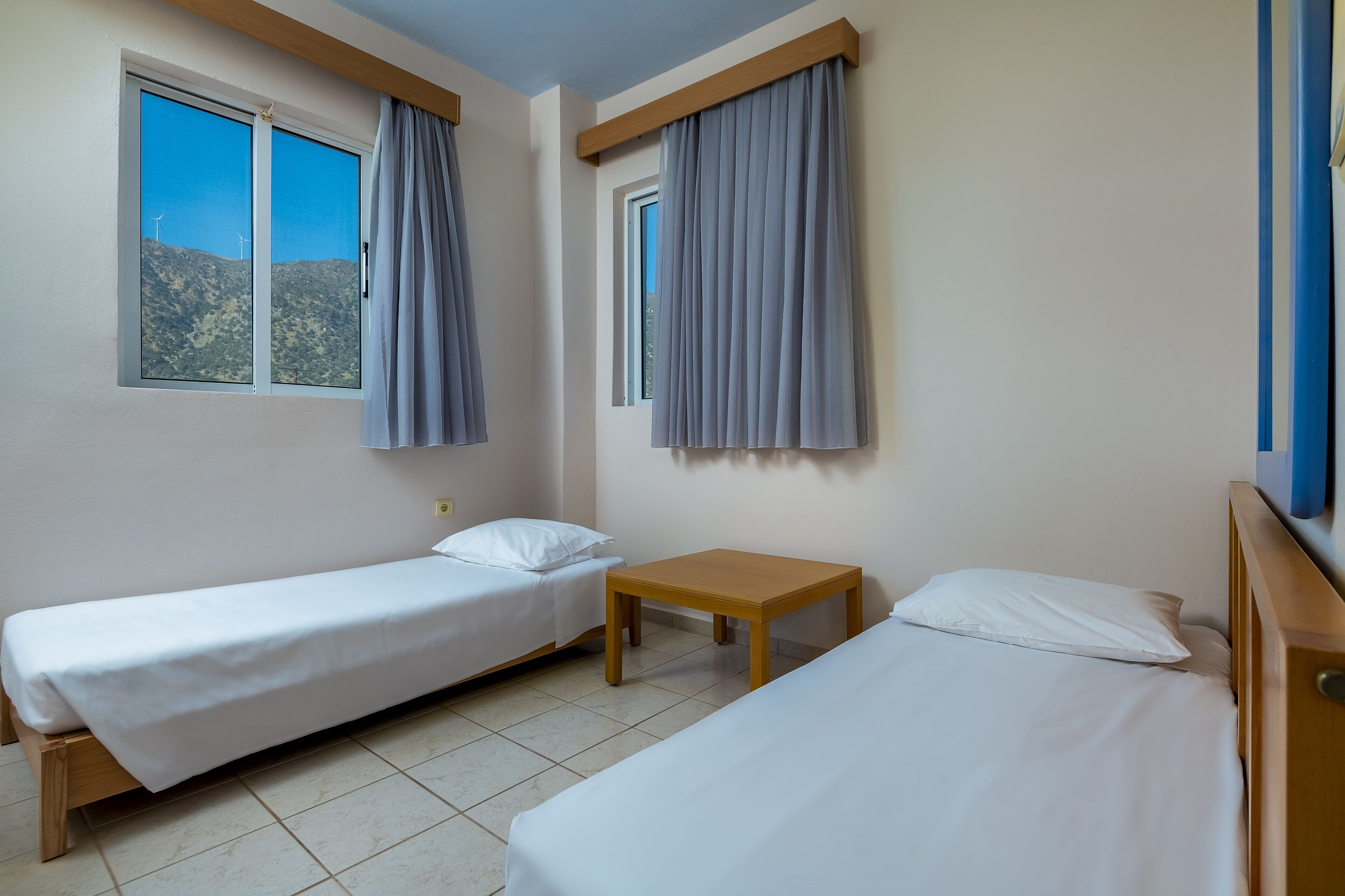 Una habitación con dos camas individuales, una mesita y dos ventanas con cortinas.