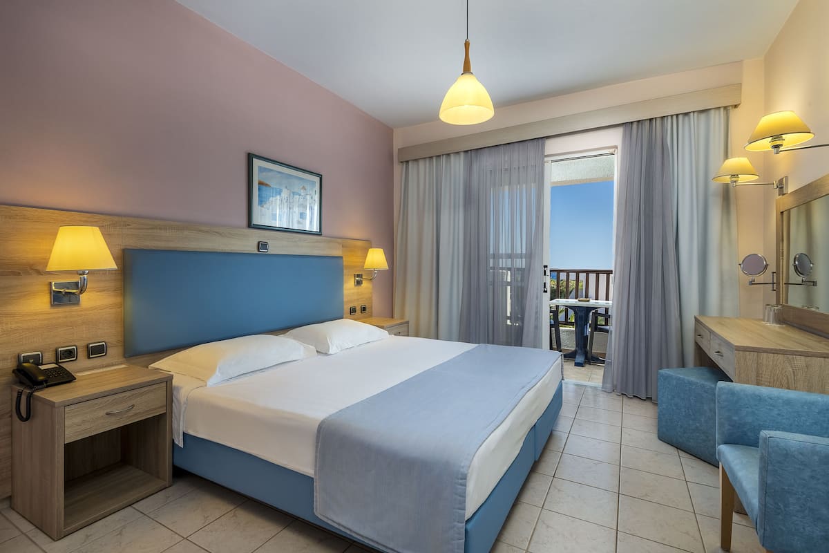 A hotel room with a large bed, bedside lamps, a desk with a chair, a balcony, and a bathroom with a mirror and towel rack.