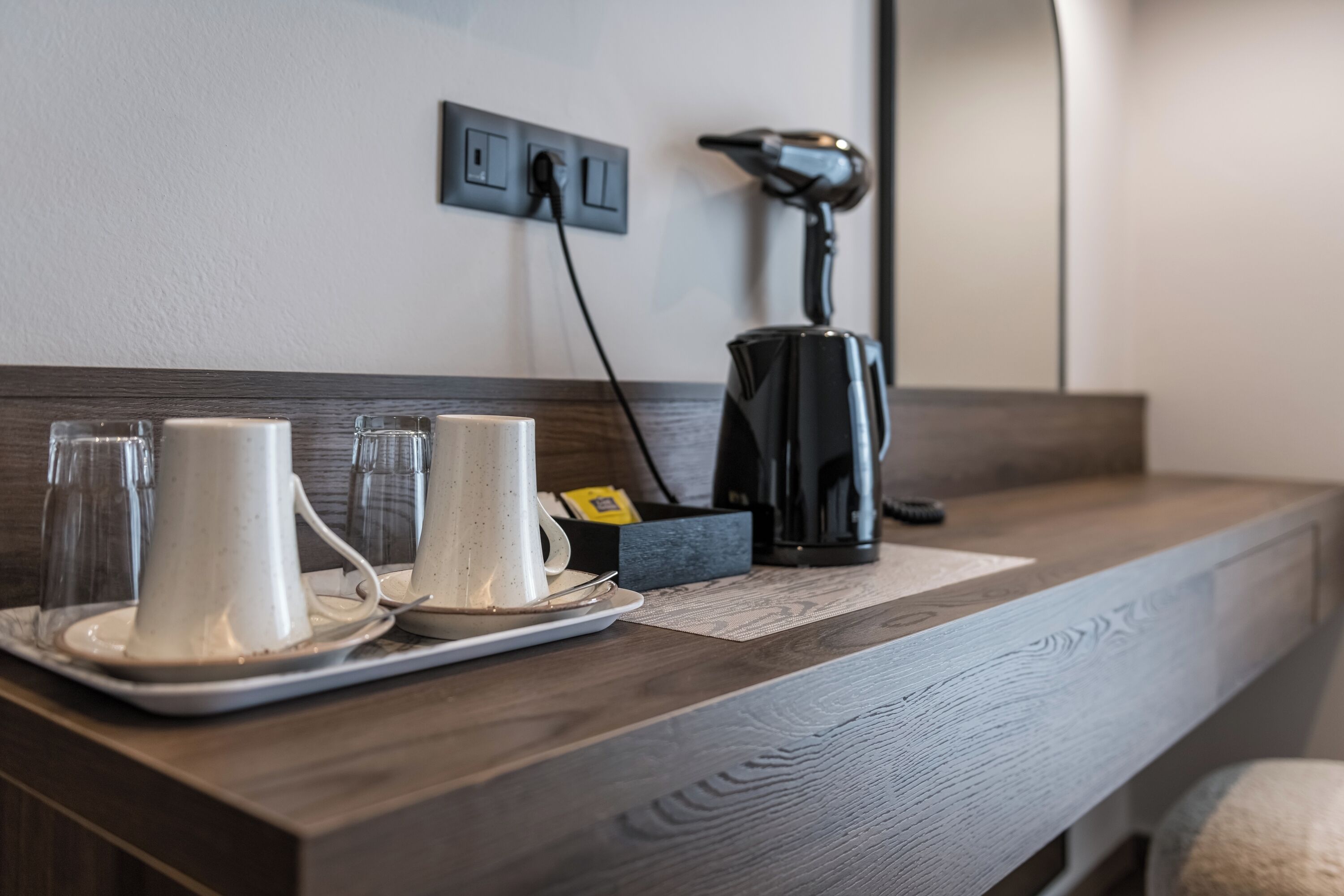 Una camera d'albergo con un comodino in legno, una lampada nera, una macchina per il caffè e un vassoio con due tazze e bicchieri bianchi.