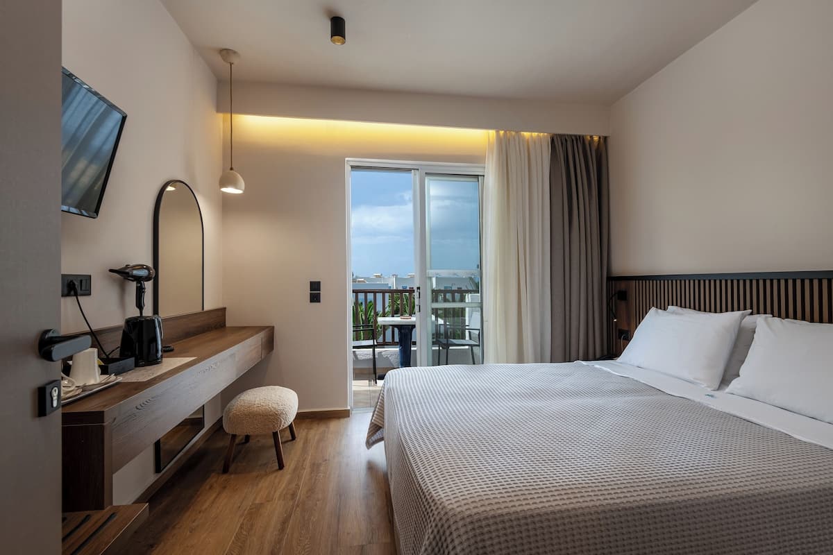 A modern hotel room with a large bed, a desk with a computer, a flat-screen TV, and a balcony with a view.