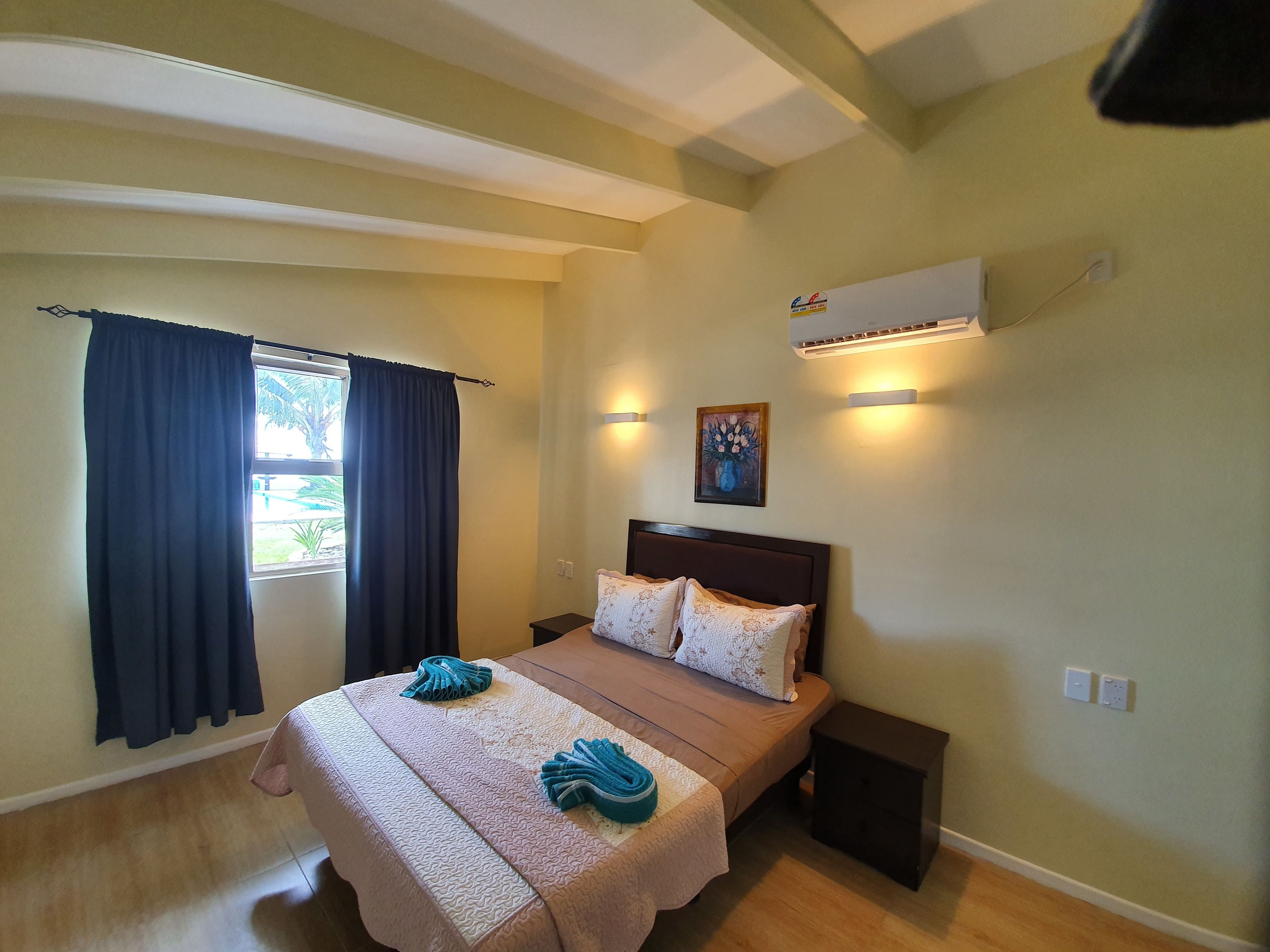 A hotel room with a bed, two bedside tables, a window with curtains, an air conditioning unit, and a painting on the wall.
