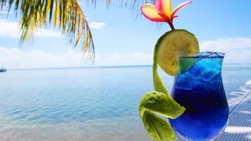 A tropical drink with a slice of lime and a flower garnish, set against a beach backdrop with clear blue water and palm trees.