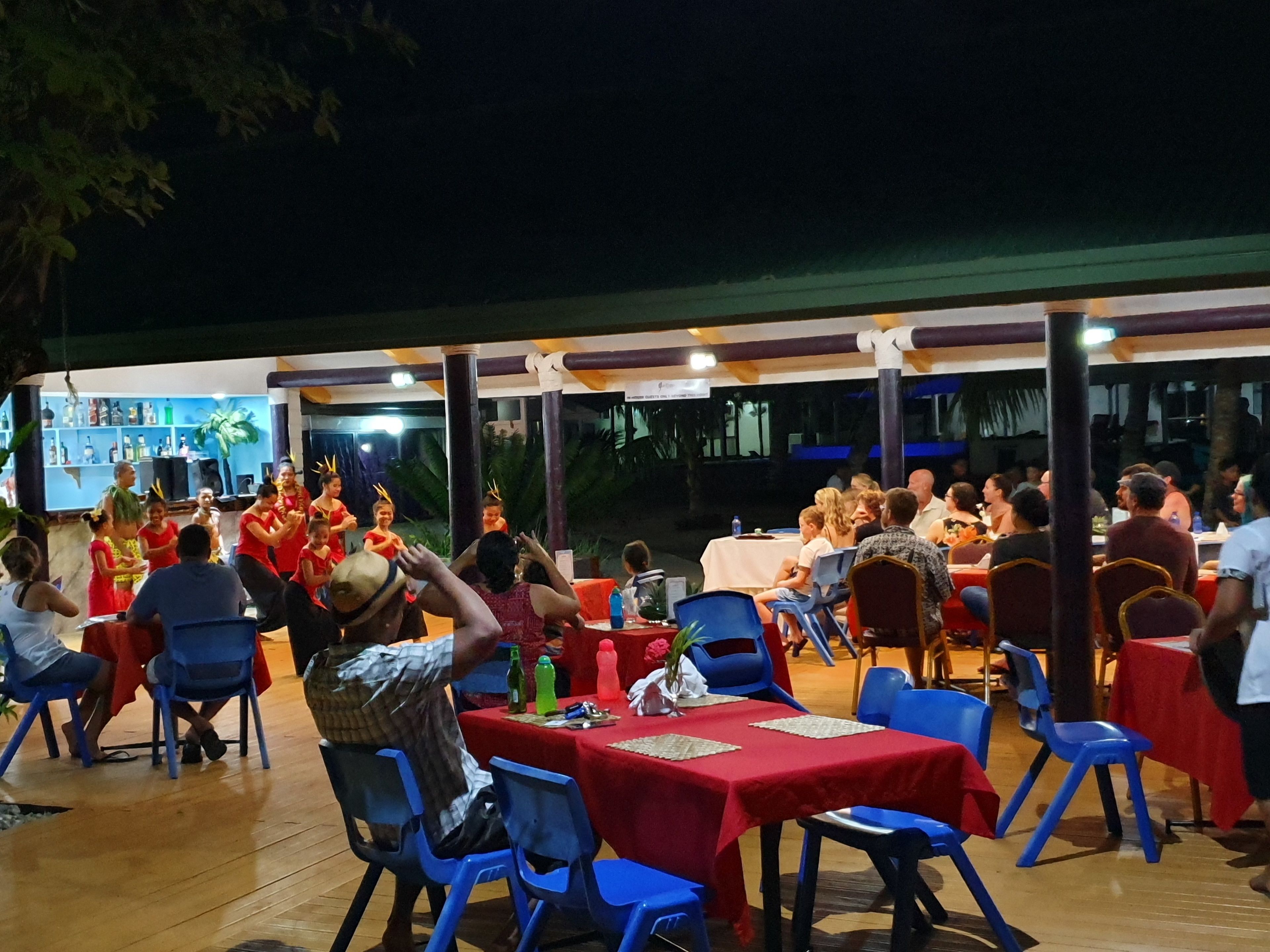 Un espacio de comedor al aire libre con mesas cubiertas de manteles rojos, sillas azules y comensales sentados.