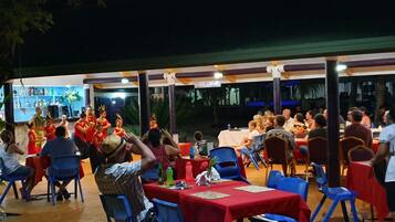 Un espacio de comedor al aire libre con mesas cubiertas de manteles rojos, sillas azules y comensales sentados.
