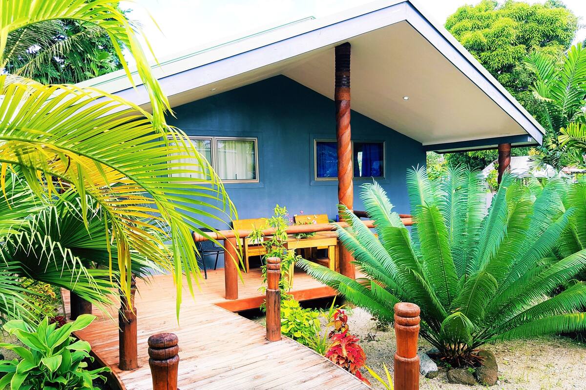 Una casa con terraza de madera, un comedor con mesa y sillas, y abundante vegetación que incluye palmeras y helechos.