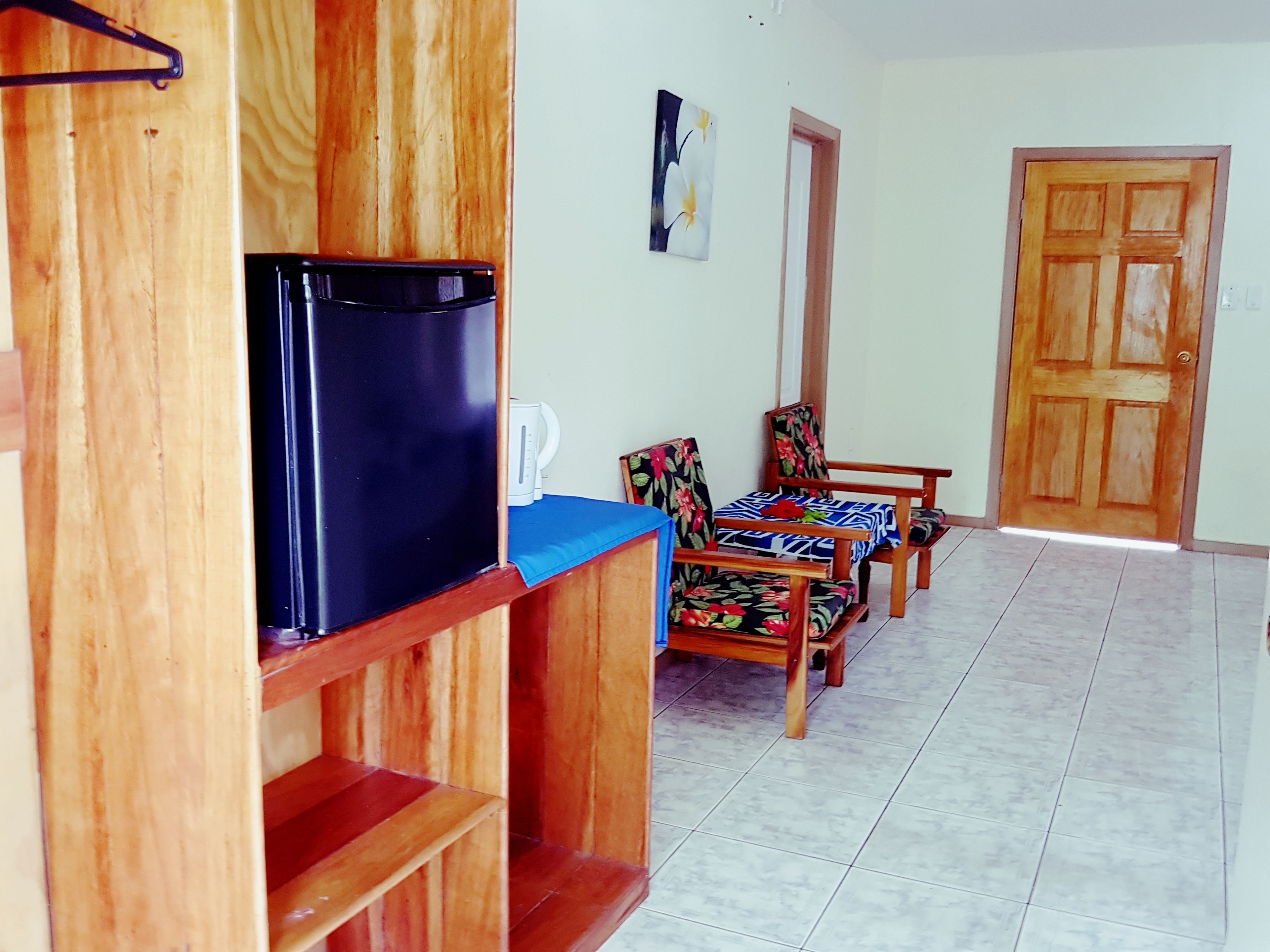 A room with a wooden cabinet, a black refrigerator, a blue table, two floral-patterned chairs, and a wooden door.