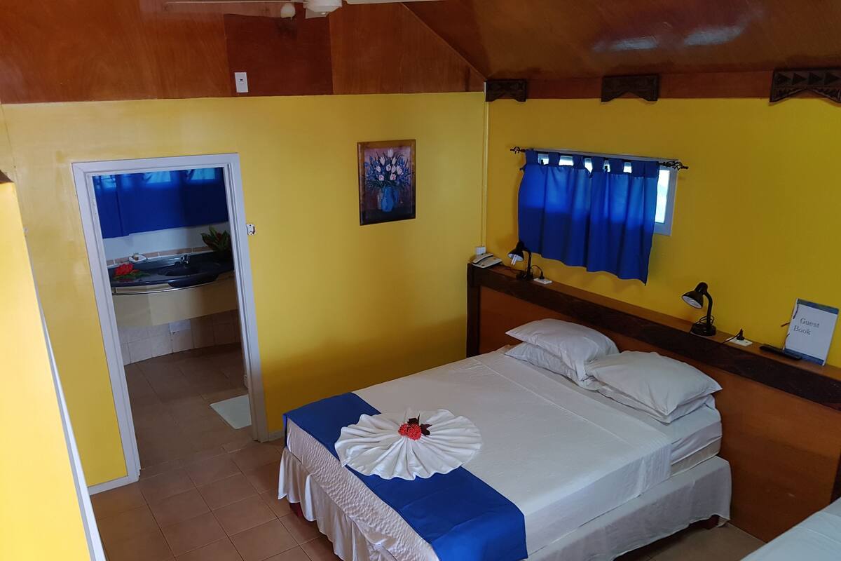 Una habitación de hotel con una cama, una mesita de noche, un escritorio, una silla, una ventana con cortinas, un cuadro en la pared y una puerta que da a otra habitación.