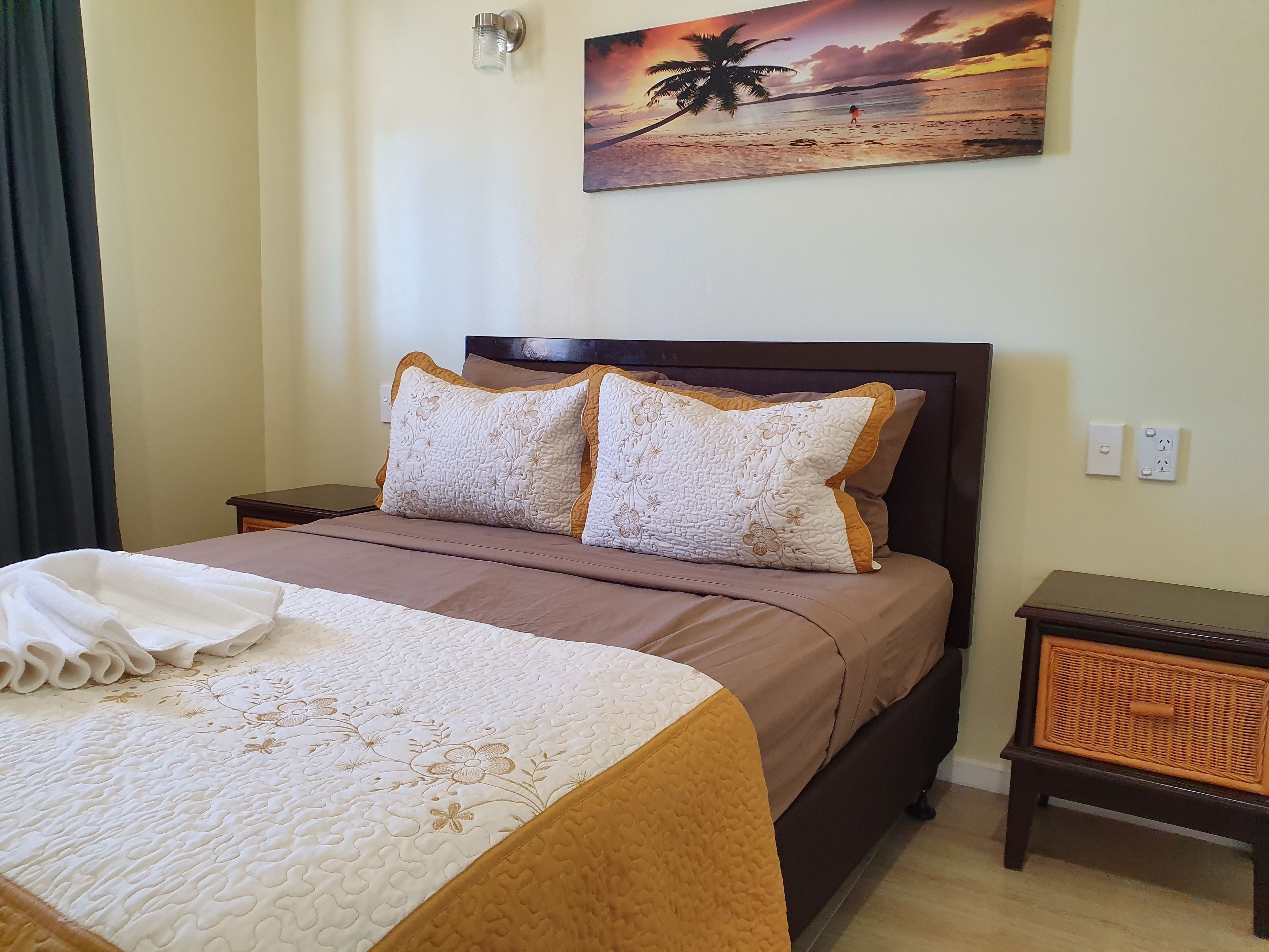 A neatly made bed with a floral-patterned bedspread and pillows, a painting of a beach scene on the wall, and a small wooden nightstand with a woven basket.