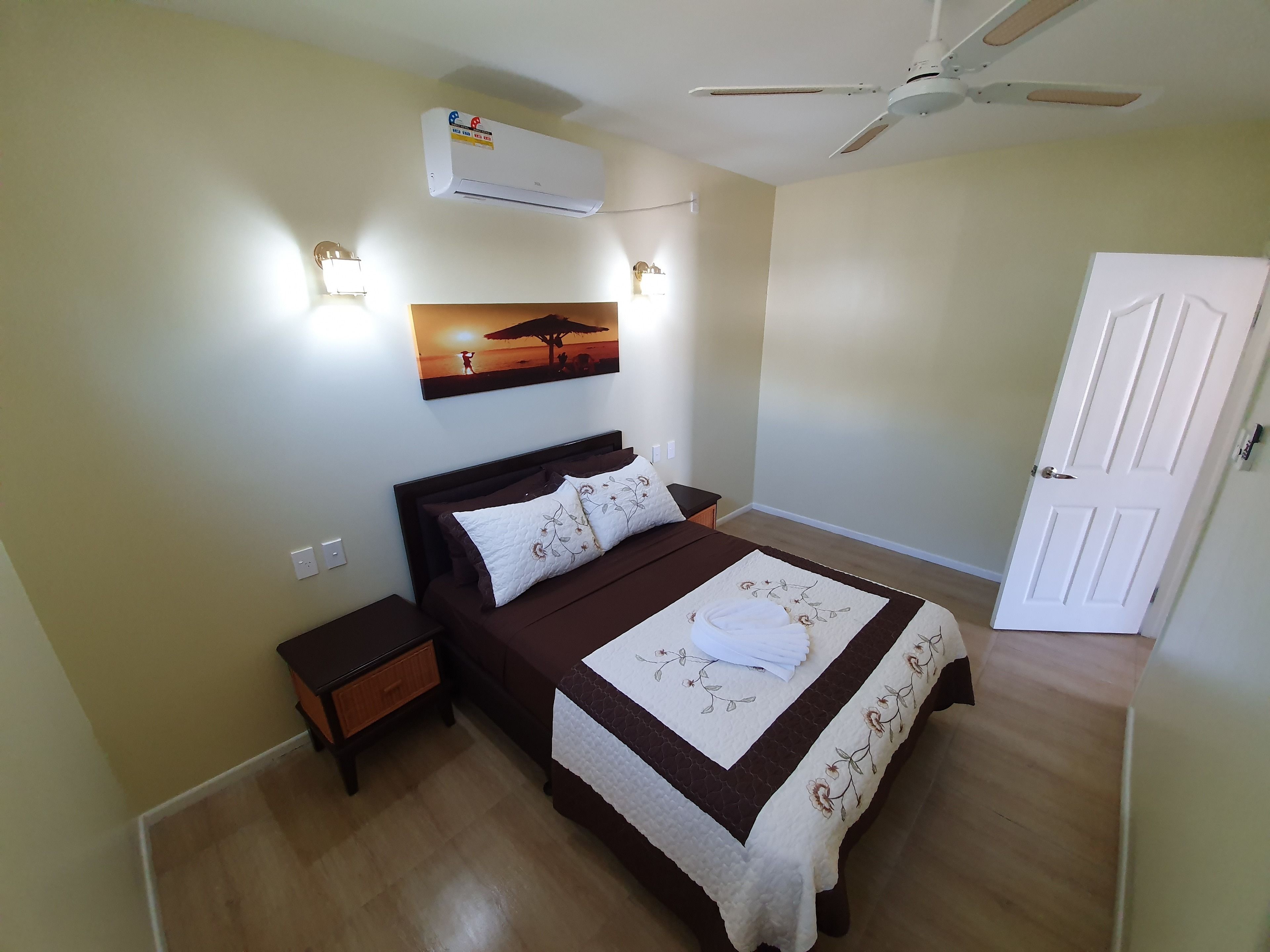 Un dormitorio con cama, mesita de noche, aire acondicionado de pared y ventilador de techo, además de una puerta.
