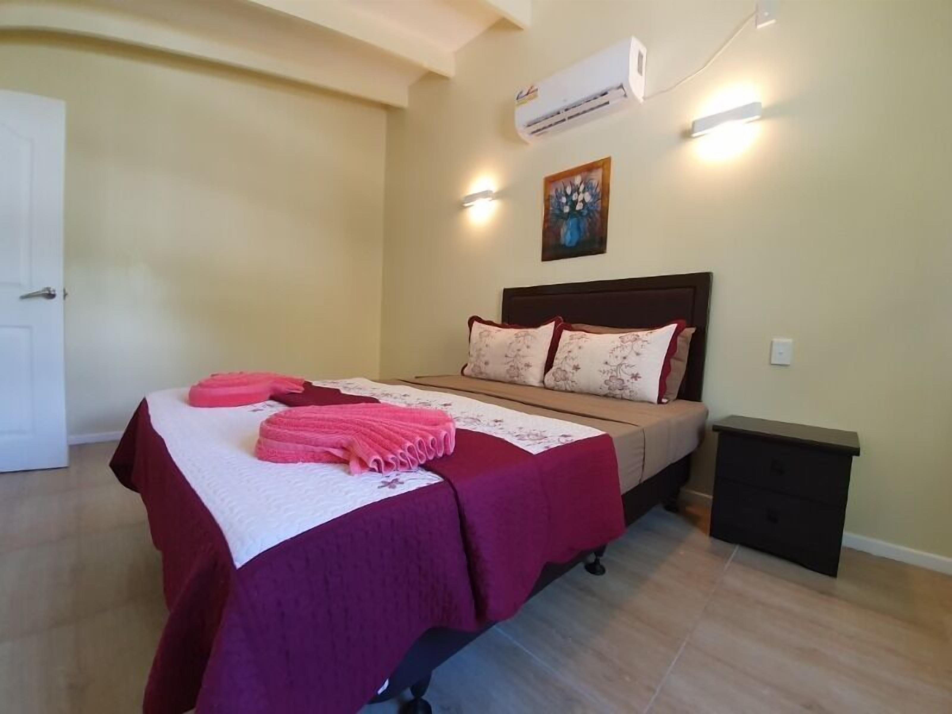 A bedroom with a bed, pillows, a bedside table, an air conditioner, and a painting on the wall.