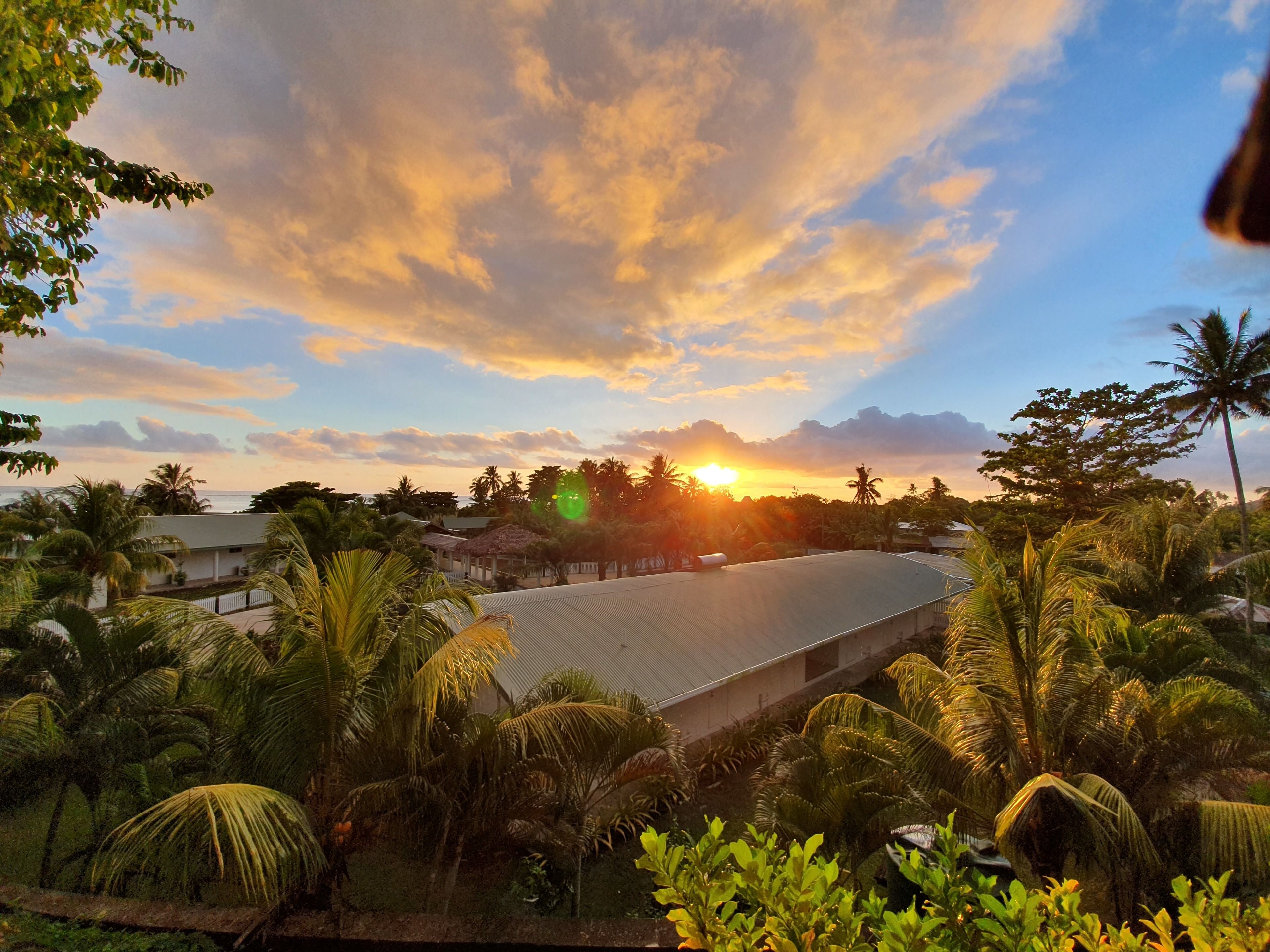 A tropical landscape with a large building, palm trees, and a sunset.