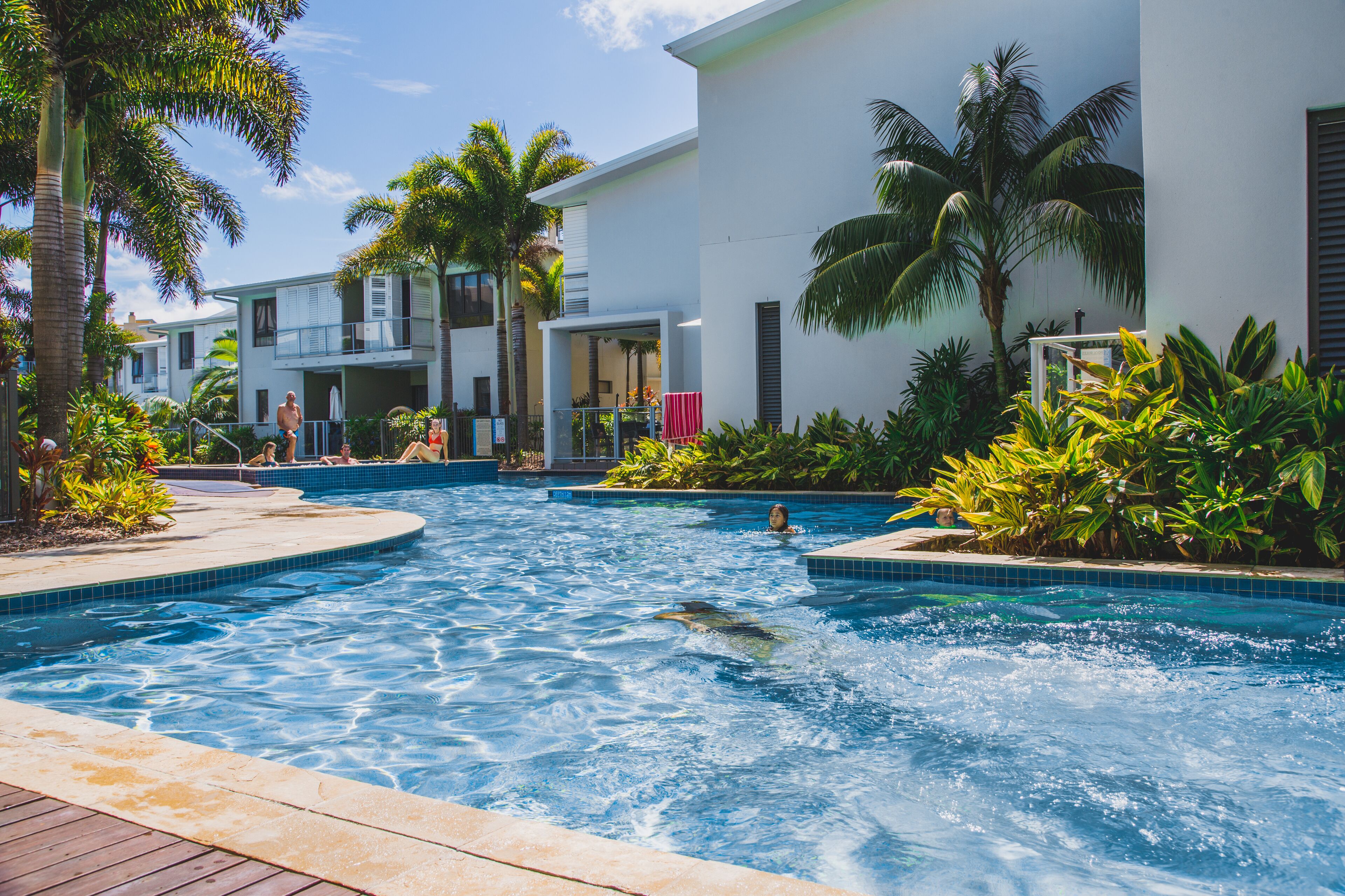 A swimming pool area with a clear view of the pool, surrounding palm trees, and a building with a balcony.