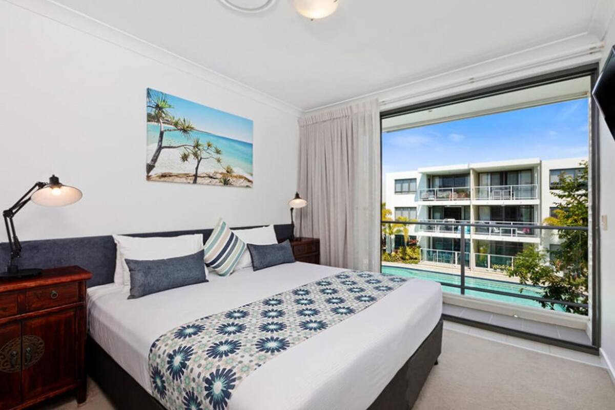 Una habitación de hotel con una cama, un cuadro de una playa, un ventanal con vistas a una piscina y edificios, y una mesita de noche con una lámpara.