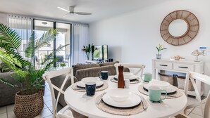Un comedor con una mesa redonda dispuesta para cuatro personas, rodeada de sillas. Se observa un televisor sobre un mueble en el fondo y un armario blanco con cajones a la derecha.