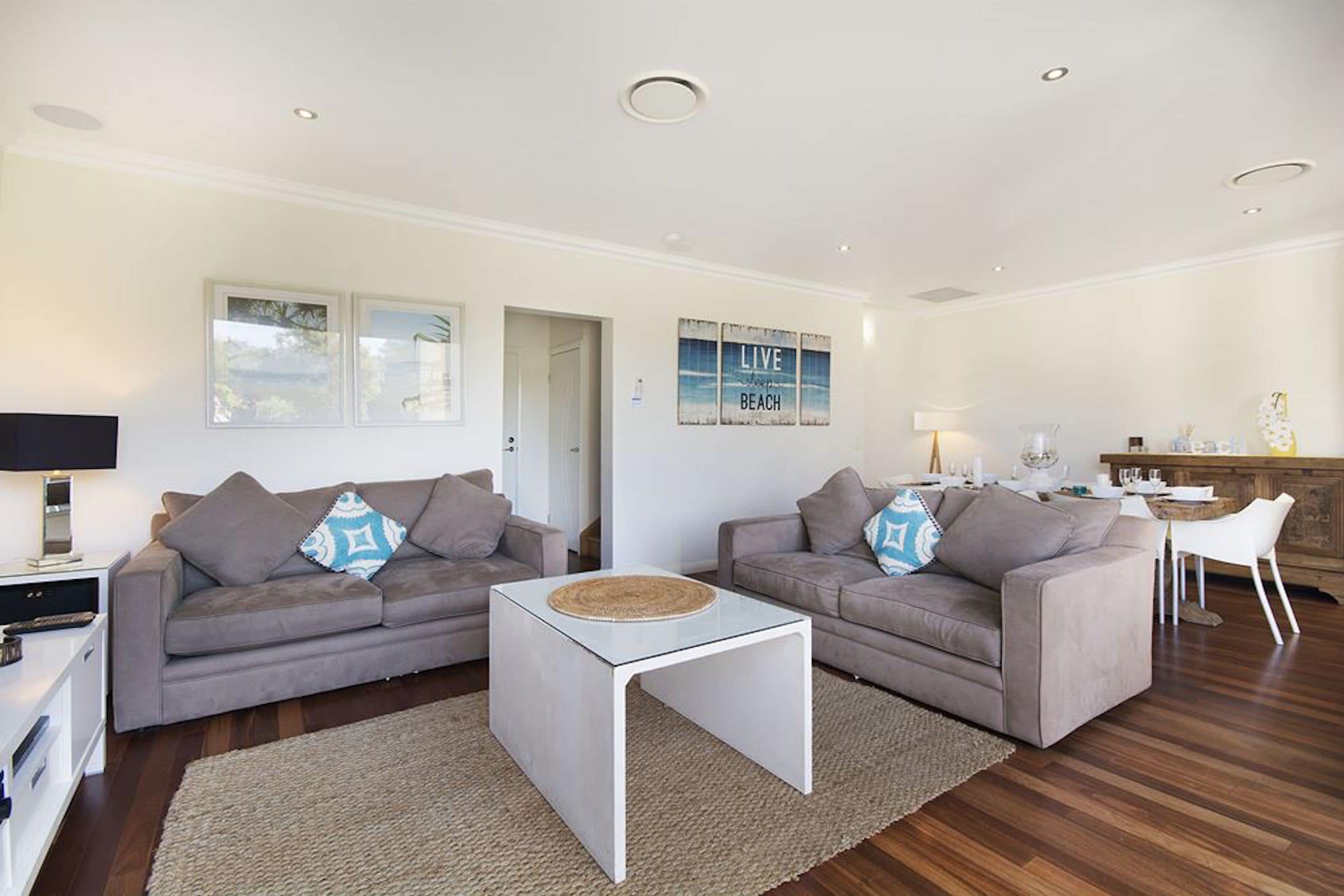 A modern living room with two grey sofas, a white coffee table, and a kitchen area in the background.