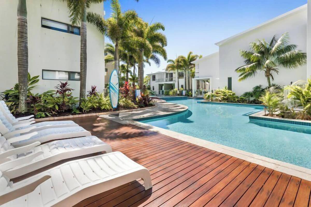 Un área de piscina con sillas de descanso, palmeras y una terraza de madera.