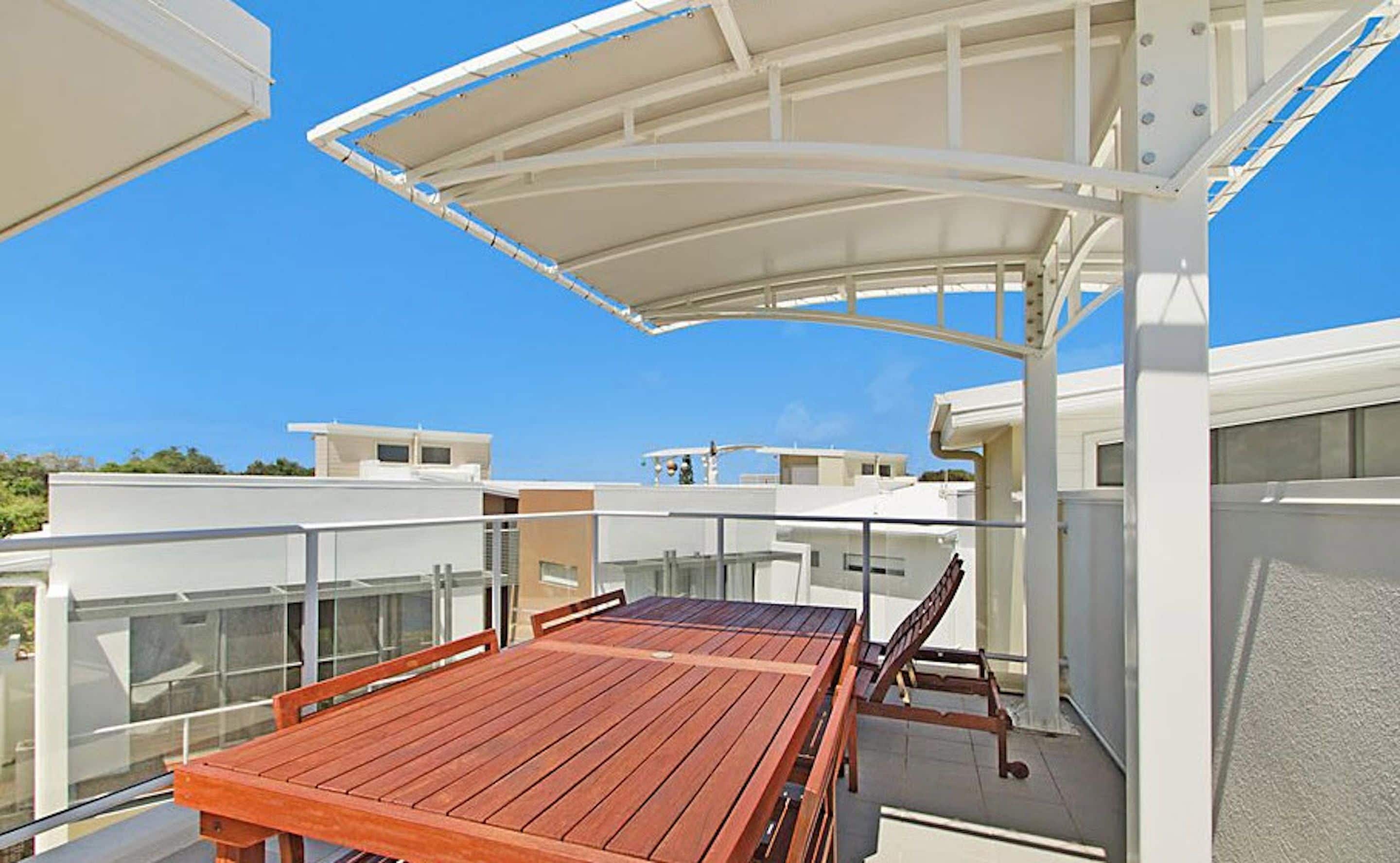 Eine moderne Terrasse im Freien mit einem Holztisch, Stühlen, einem weißen Sonnendach und freiem Blick auf die umliegenden Gebäude.