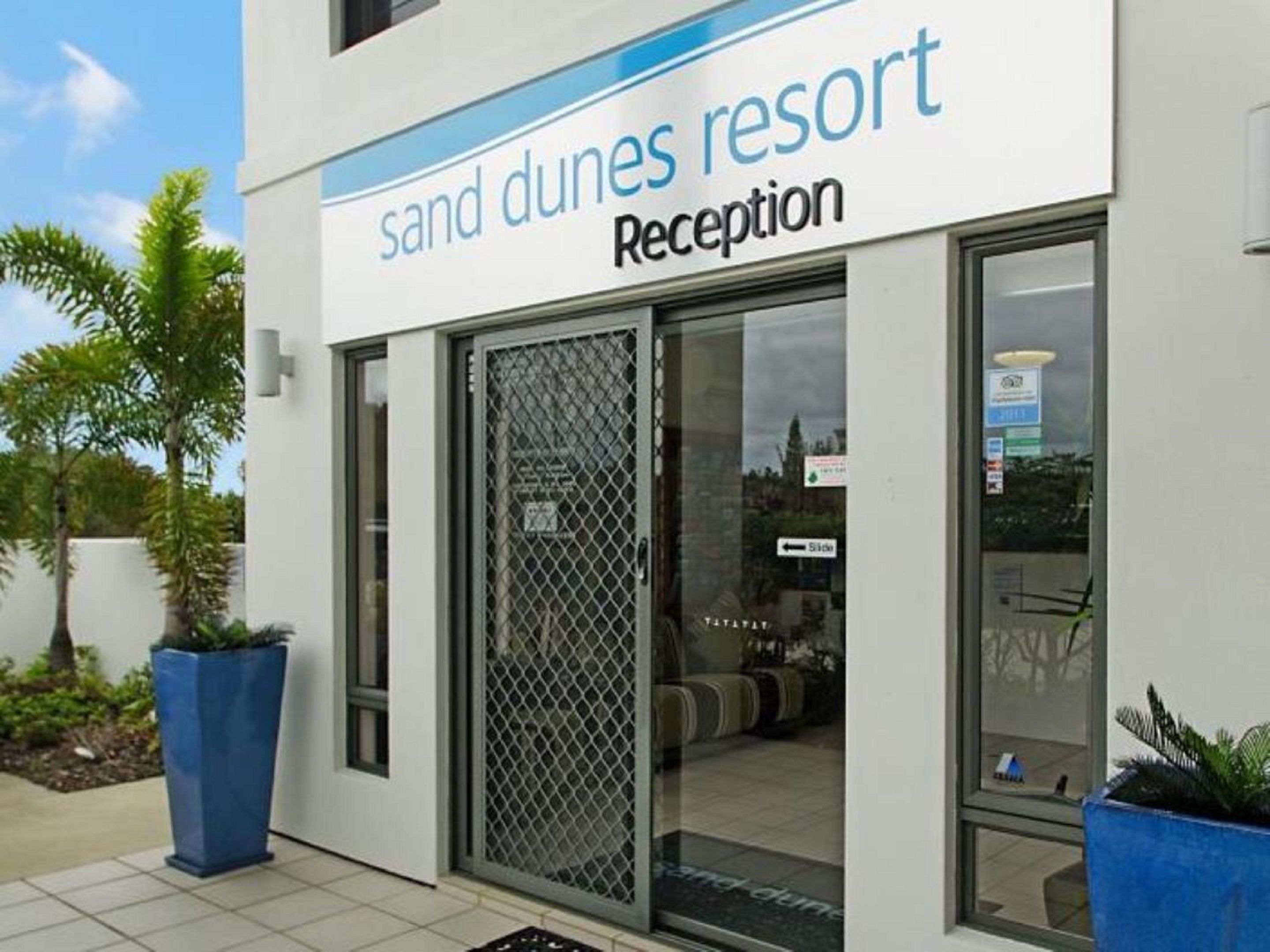 Un espace d’accueil à Sand Dunes Resort, avec une porte en verre transparent, une enseigne au-dessus indiquant la réception, et des plantes en pot à l’extérieur.