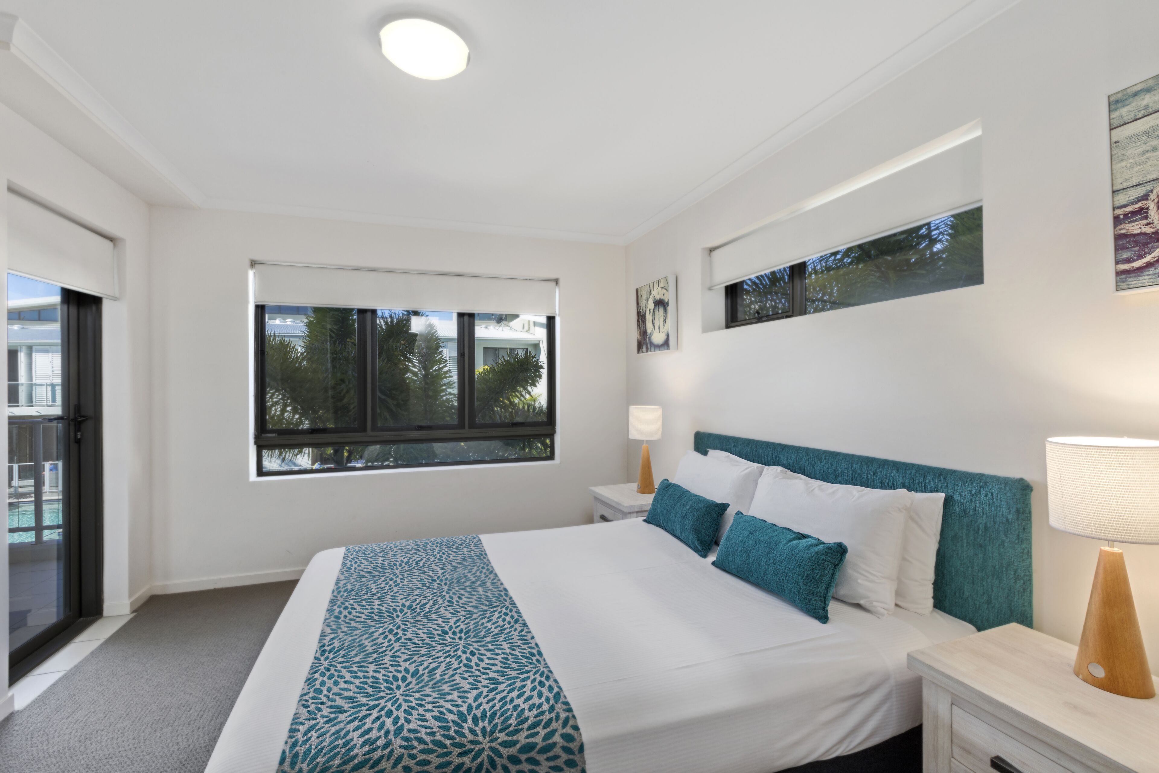 Une chambre moderne avec un grand lit, une fenêtre donnant sur des palmiers, une table de chevet avec une lampe et un luminaire au plafond.